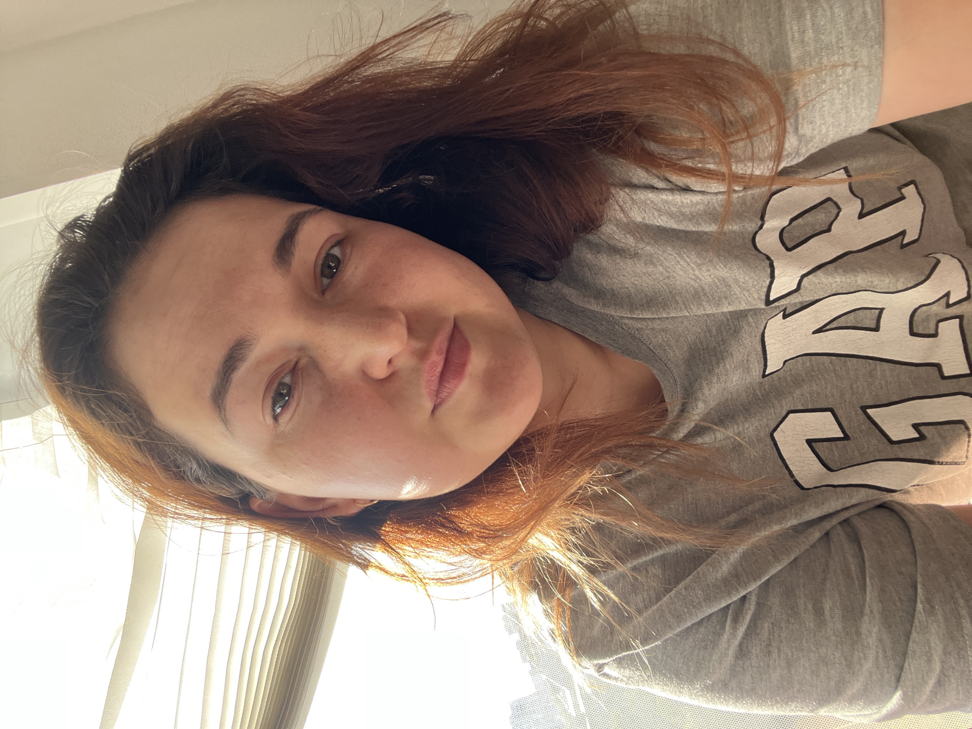 This is a selfie of a woman with reddish-brown hair and light skin, with a smattering of freckles across her nose and cheeks. She is wearing a grey t-shirt with black lettering that appears to spell out "FREE". Her expression is neutral, and she is looking directly at the camera. The lighting suggests it is daytime, with sunlight streaming in from the left side of the frame, creating some shadows on her face. The background is slightly out of focus but appears to be an indoor setting with vertical blinds visible on the left. The image is oriented sideways.