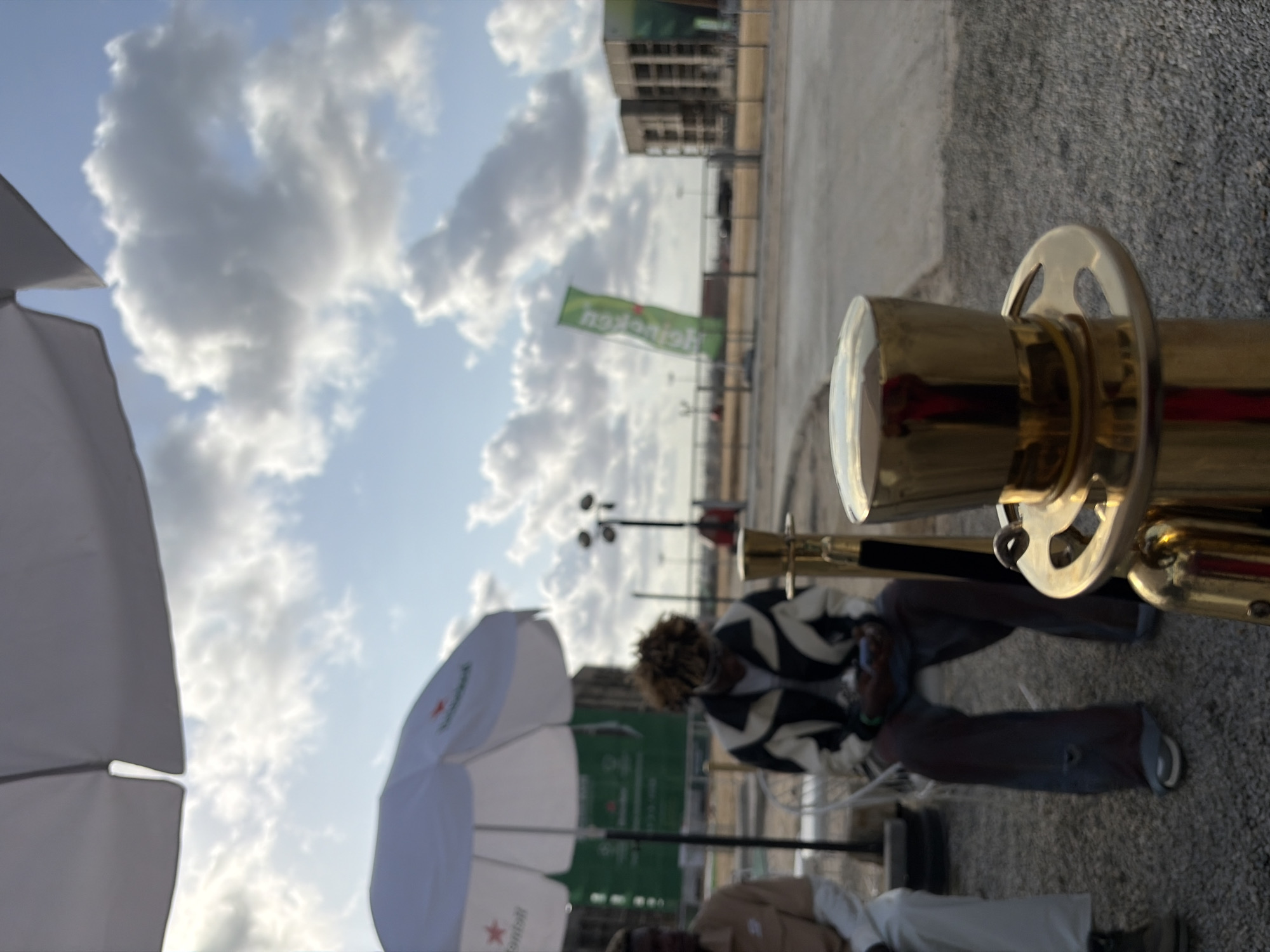 The image, likely taken in Lagos, Nigeria, presents a blurred scene with a mix of foreground and background elements.
In the foreground, a gold-colored stanchion post dominates the right side of the frame, possibly used for crowd control or VIP area demarcation.
Behind this, we can see a person wearing a white and black patterned jacket sitting at a table. A white umbrella with a Heineken logo is visible, suggesting a sponsored or branded event.
Another person is partially visible, dressed in khaki or beige clothing, sitting at the same table.
In the background, there are structures that seem like buildings, along with what appears to be a Heineken flag. The sky is partly cloudy. The picture appears to be taken during the day.