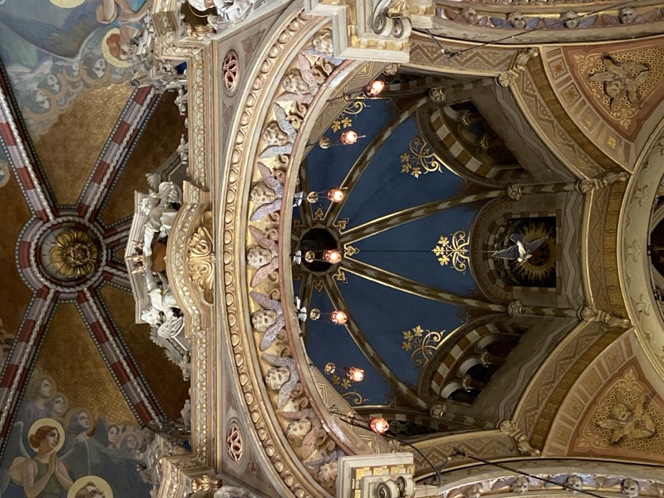 An ornately decorated ceiling features gold and blue patterns emanating from a central point. Intricate molding with cherubic figures lines the edges of the dome-like structure. A gilded flower design is visible on a neighboring ceiling section. Hanging lights are suspended from the center, and a bird statue adorns one of the panels. The overall aesthetic suggests a religious or historical setting.