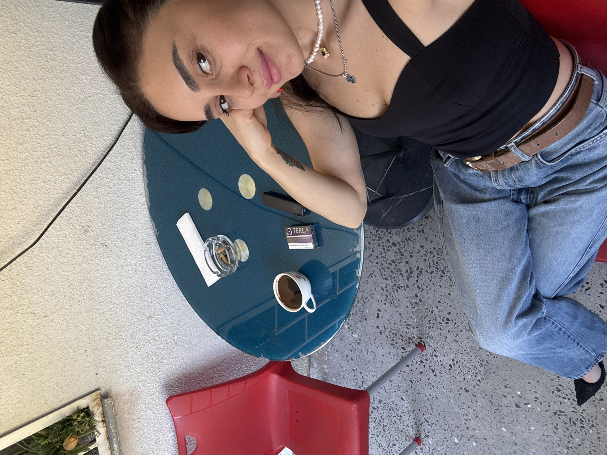 A woman is sitting at a round, blue table outdoors, with a red chair to her left. She is wearing a black top, blue jeans, and black shoes. On the table, there is a cup of coffee, an ashtray with cigarette butts, a pack of cigarettes, and a white napkin. The background shows a textured wall and the ground is a speckled concrete. The scene is likely on a patio or balcony in Tbilisi, Georgia.