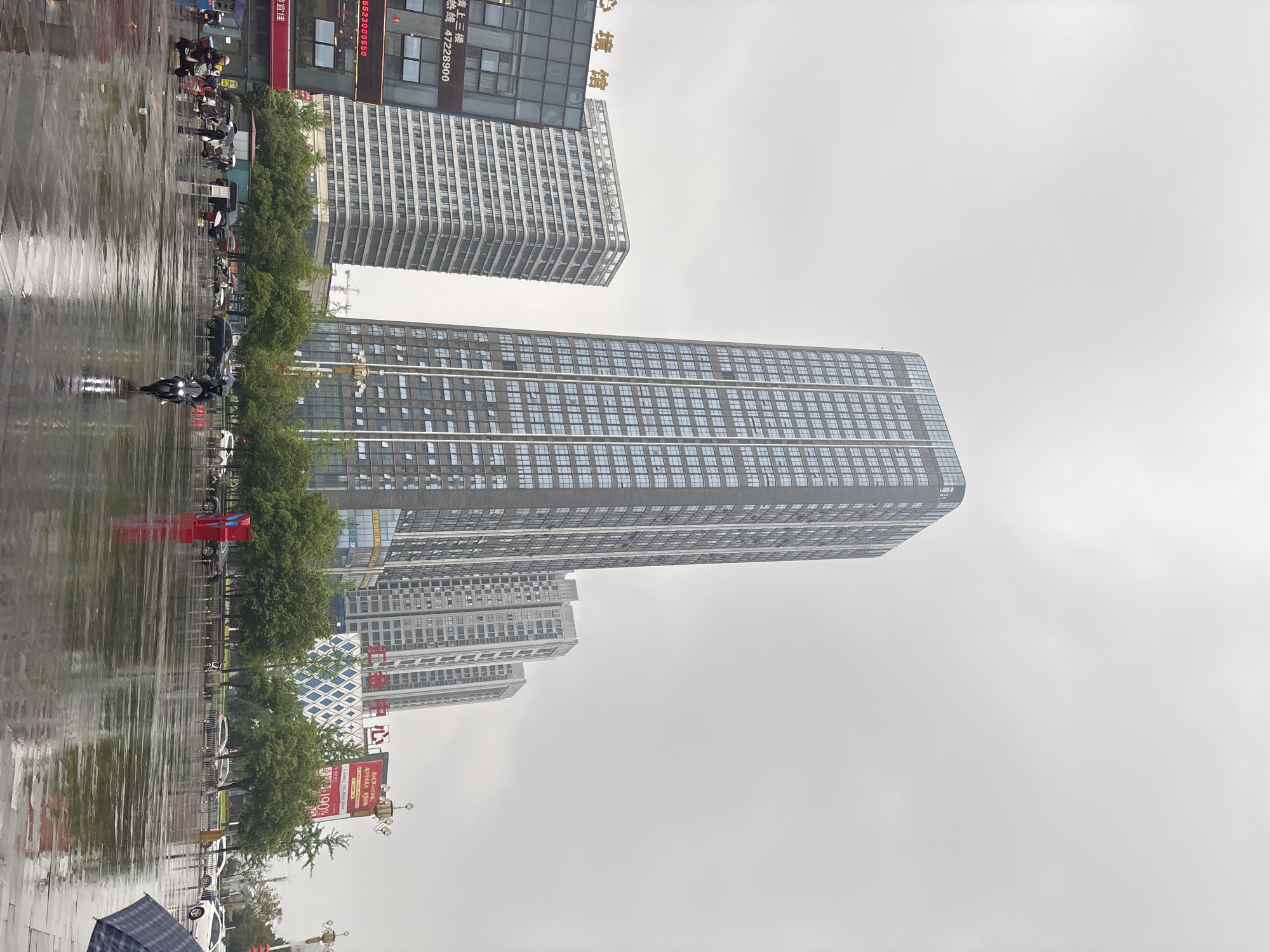 The image depicts a street scene in Jiangjin, China, on a rainy day. The foreground features a wet pavement reflecting the surrounding buildings and street activity. Trees line the street, separating the pavement from the road.
Multiple high-rise buildings dominate the background, with one particularly tall skyscraper centrally located. The architecture is modern, with many windows visible on each building.
People are present in the scene, primarily traveling on motorcycles or walking with umbrellas. The sky is overcast and grey, indicative of rain. Street signs and commercial advertisements are visible.