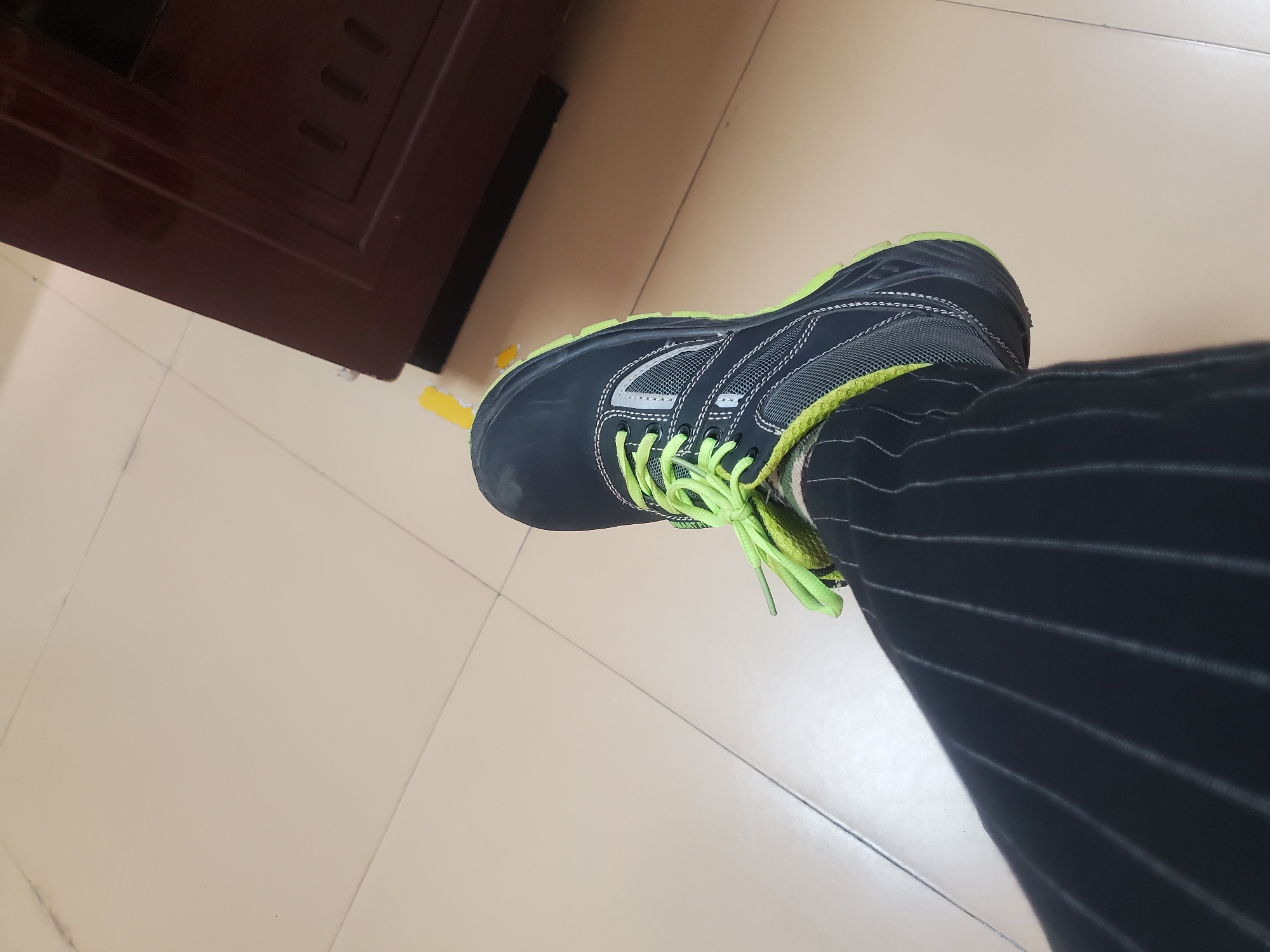 The image depicts a black and grey running shoe with bright green laces and sole trim, resting on a beige tiled floor. The shoe is on a person's foot, which is covered by black pinstripe pants. A dark brown cabinet is partially visible in the upper left corner of the image. The setting appears to be indoors on a light tile flooring. There is no evidence to suggest that the image was taken in Gwagwada, Nigeria.