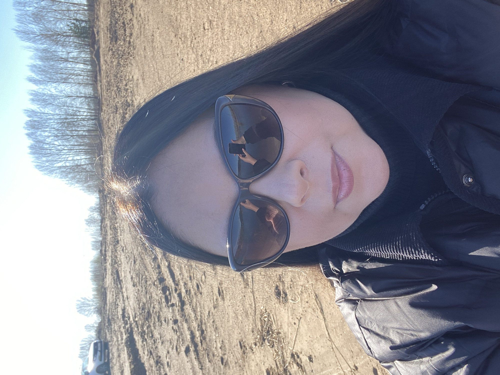 A woman is pictured outdoors in Noviy, Russia, as evidenced by the trees visible. She is wearing sunglasses and a black jacket. The ground appears to be dry and possibly agricultural. The reflection in her sunglasses shows her holding a mobile phone, suggesting she is taking the photo.
