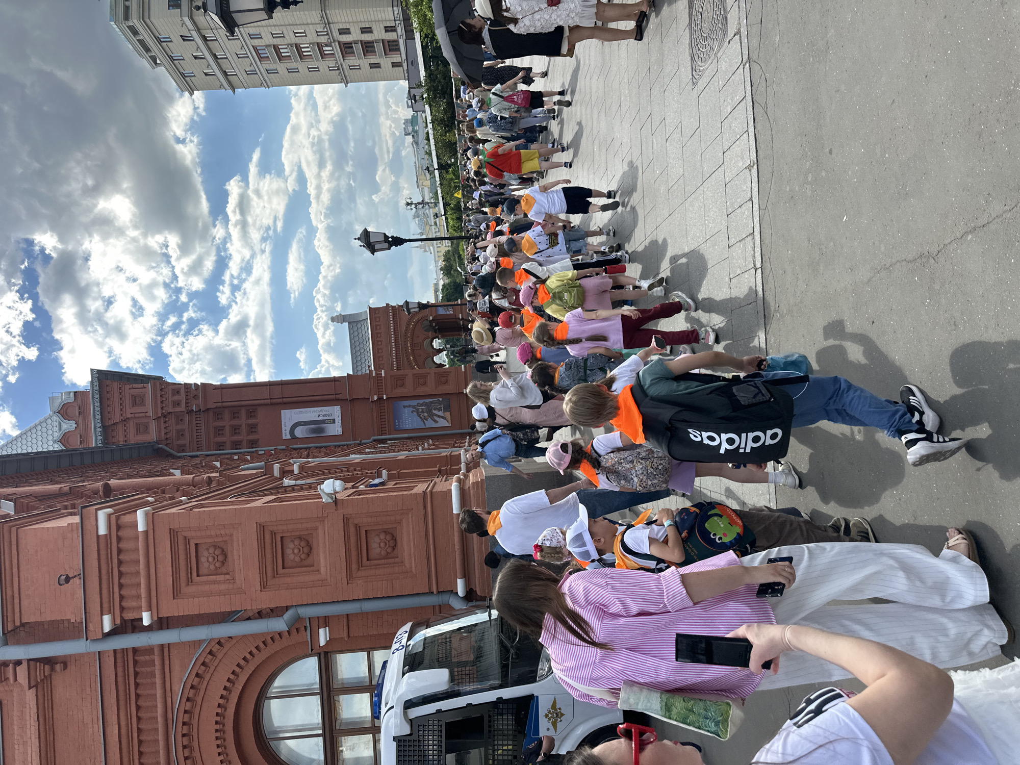The image depicts a lively street scene in Moscow, Russia, captured from a slightly elevated vantage point. A large group of people, primarily children and young adults, is walking along a broad sidewalk. Many of the children are wearing bright orange or yellow sashes or vests, indicating they are part of an organized group, possibly a tour or school excursion. Several adults accompany them, some holding umbrellas.
The setting features prominent architecture on both sides of the street. On the left, a grand reddish-brown building with intricate brickwork, decorative elements, and arched windows lines the sidewalk. Partially visible text on a banner or sign on this building reads "СВОБОДА" (Freedom) and possibly "ПАЛЬМЫ И ЦВЕТЫ" (Palms and Flowers). On the right, another building with multiple windows and a different facade is visible.
The scene unfolds under a partly cloudy blue sky, suggesting a sunny daytime. Shadows on the ground indicate bright light, likely late morning or early afternoon. A white van, identified as a police vehicle by the text "Пoлиция" (Police) on its side, is visible near the group. Other notable objects include a black backpack with the "adidas" logo worn by one person. The ground transitions from paved asphalt to tiled or cobblestone sections. The overall impression is one of an active, well-attended public outing or tour.