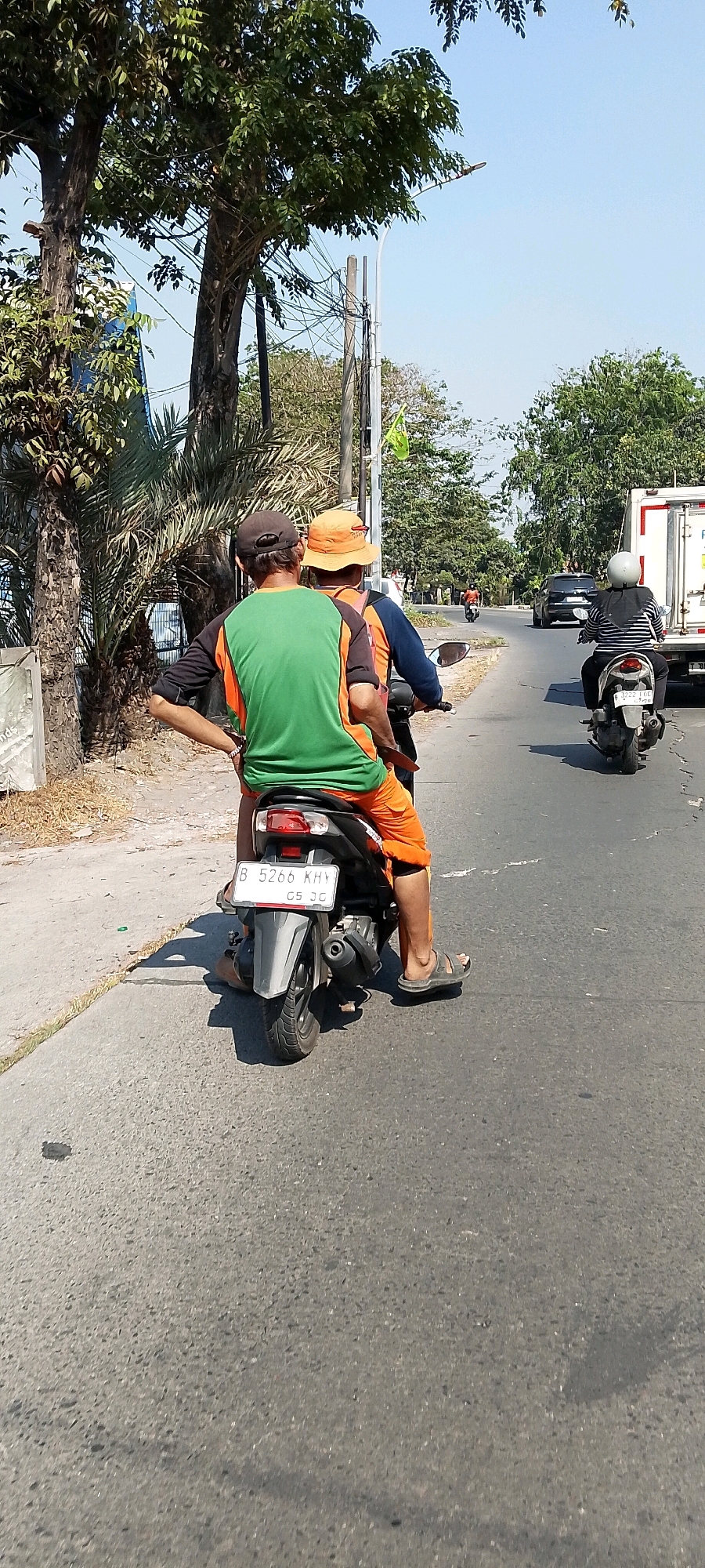 In the city of Bekasi, Indonesia, two men are riding a motorcycle down a paved road on a sunny day. The driver, wearing an orange hat and a dark blue shirt, is on the front. Behind him, the passenger wears a dark baseball cap, a green t-shirt with orange trim, and orange shorts. Both are facing away from the camera. The motorcycle has a license plate that reads "B 5266 KHY CS JG". On the right side of the road, another motorcycle carries two people, and a white truck with a box trailer is also visible. The environment includes trees and a clear blue sky.