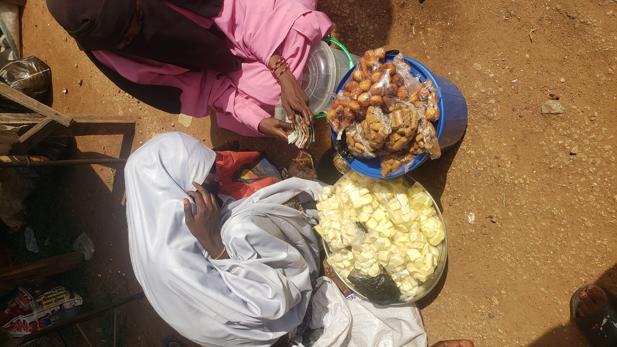 Two people are engaged in a transaction at an outdoor market stall in Kakuri Gwari, Nigeria. One individual, wearing a pink hijab and a pink garment, is handing money to the other, who is draped in a white hijab and a white garment. The person in pink appears to be a vendor, as they are seated next to two buckets overflowing with goods. One bucket contains numerous small, golden-brown fried snacks, likely sweet, packaged in clear plastic. The second bucket holds a pile of yellow, cubed items, also in plastic. To the lower right, another person's feet are visible in sandals, suggesting they may be a customer. The ground is dirt, typical of an outdoor market. The sunlight casts distinct shadows, indicating it is likely daytime. The scene appears to be a bustling, everyday market interaction.