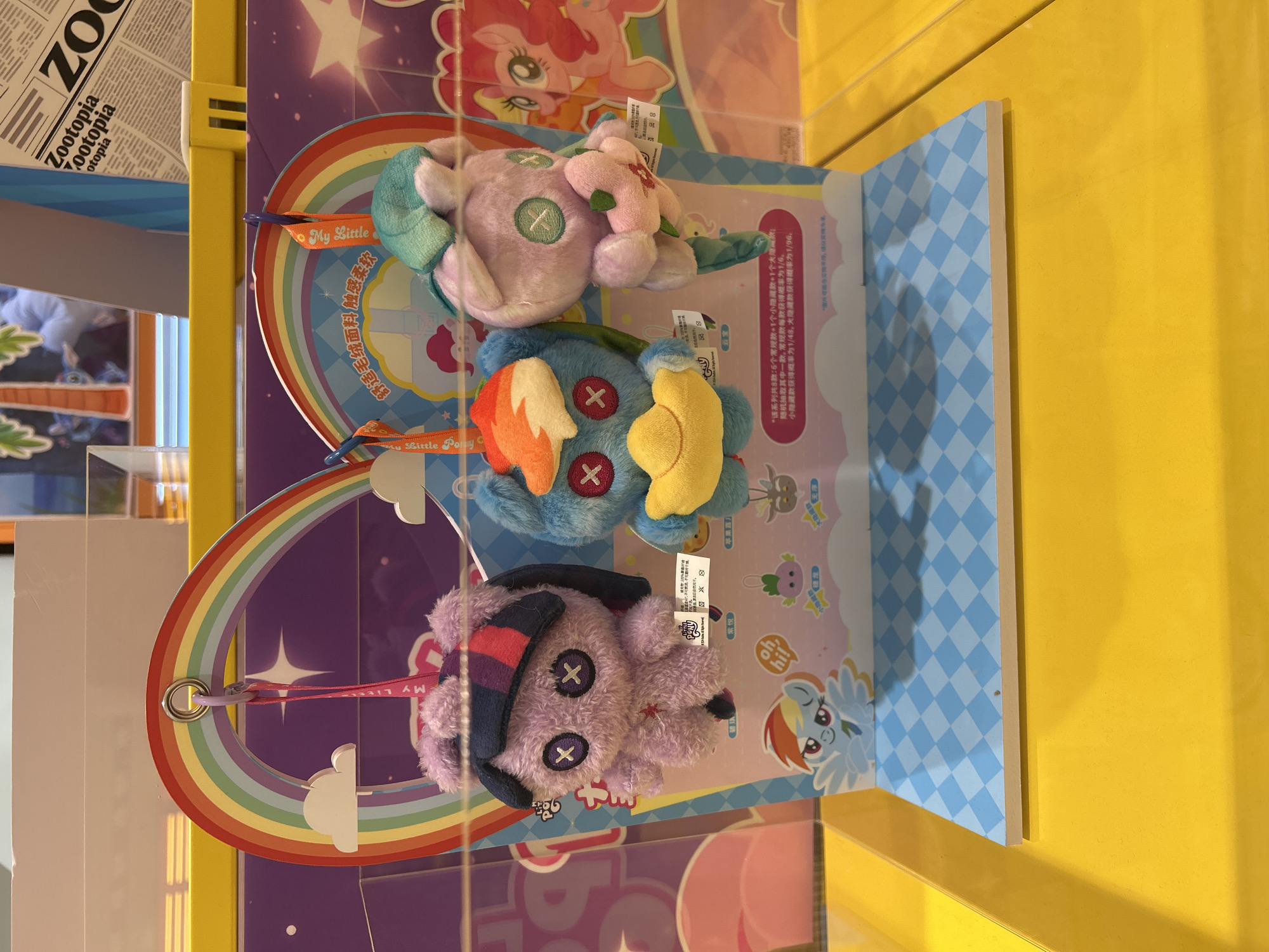 Three plush toys resembling characters from My Little Pony hang from a display stand. The toys are purple, blue, and pink, each with distinctive features like stitched-on eyes and colorful manes. The purple toy has a dark blue and purple striped headpiece. The blue toy has an orange and white mane and holds a yellow object. The pink toy has a green hat and is holding a green sprout. The display stand is adorned with a rainbow, clouds, and checkered patterns. Behind the toys, a backdrop features images of My Little Pony characters and text in Chinese. In the upper right corner, a partial view of a newspaper or magazine with the headline "Zootopia" is visible. The display is placed on a yellow surface. The image was captured in an indoor retail environment.