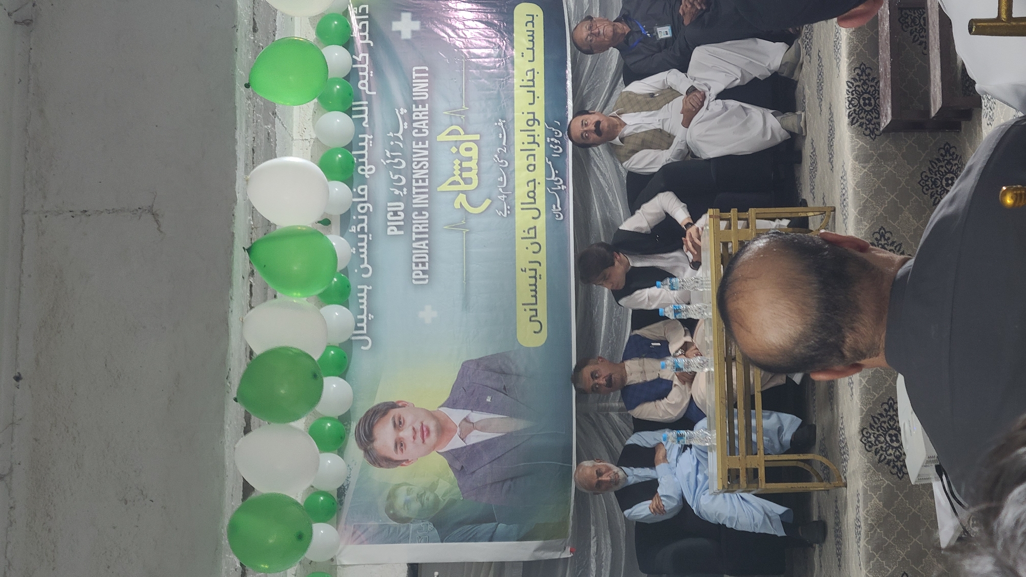 The image shows an event related to the "Dr. Kaleem Ullah Health Foundation Hospital" and the inauguration of a Pediatric Intensive Care Unit (PICU). The event is taking place in Quetta City Tehsil, Pakistan. A banner is prominently displayed with the image of a man, possibly Dr. Kaleem Ullah, and text in Urdu. The banner indicates the event is sponsored by "Nawabzada Jamal Khan Raisani, Member of the National Assembly of Pakistan." Several men are seated on chairs. There are white and green balloons decorating the space.