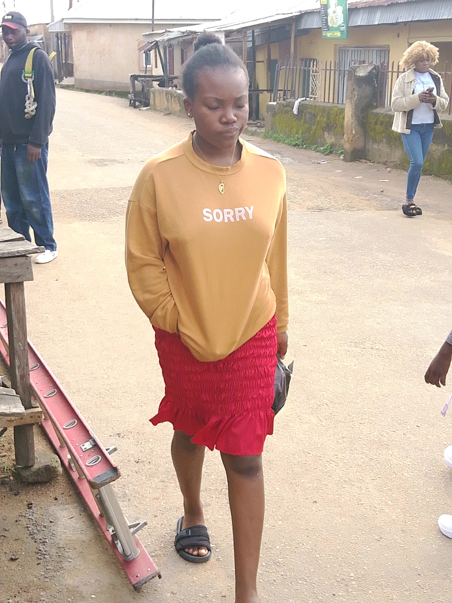 In the city of Jos, Nigeria, a young woman with dark skin and her hair in a bun walks forward. She wears a tan sweatshirt with the word "SORRY" printed in white letters across the chest and a short, ruched red skirt. A small gold necklace with a pendant hangs around her neck. She is holding a black plastic bag in her right hand. On her feet, she wears black sandals. To her left, a man in a black hooded sweatshirt and blue jeans stands with a yellow strap attached to his shoulder. He is wearing a red and black baseball cap. In the background, a woman with blonde curly hair, wearing a cream-colored jacket over a white t-shirt and blue jeans, walks away from the camera, looking at her phone. The scene appears to be a street in a residential or commercial area, with buildings lining the background and a dusty, paved road. A red ladder-like object is positioned on the left side of the frame.
