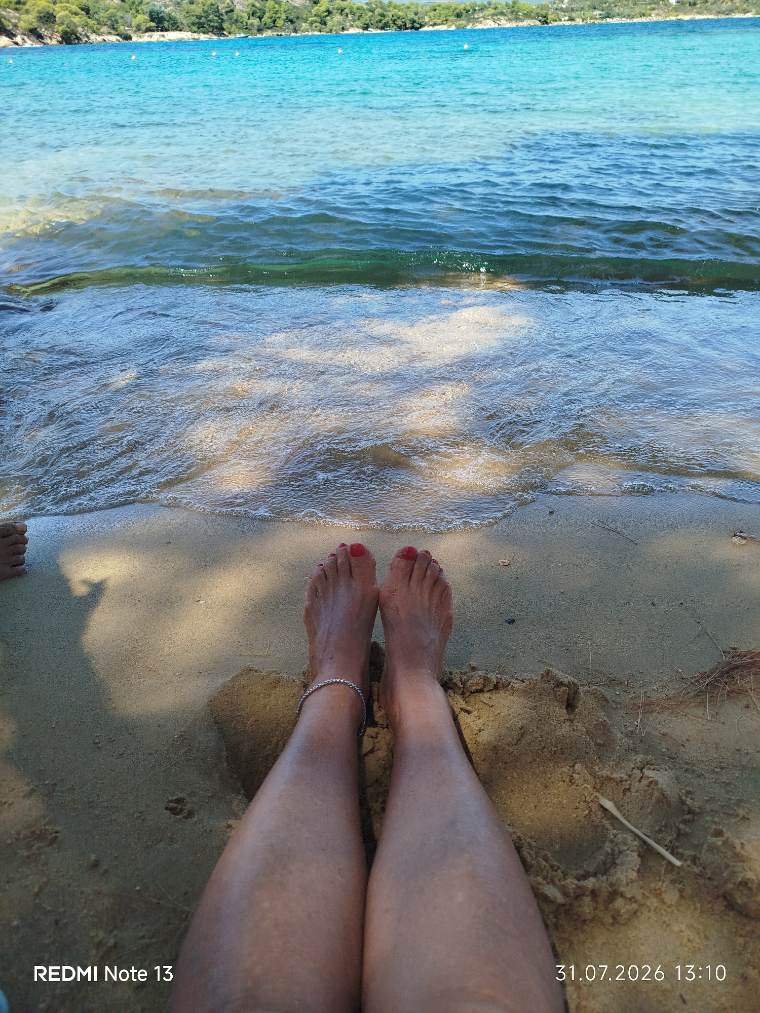 In this close-up, low-angle shot, two pairs of bare feet are positioned on a sandy beach, with the gentle waves of a turquoise sea lapping at the shore. The person on the right has red nail polish on their toes and a delicate silver anklet. The sand is light brown and appears damp near the water's edge. In the background, the clear water transitions to a deeper blue, and beyond that, a lush green coastline with trees is visible under a clear sky. The lighting suggests it is daytime, possibly late afternoon given the shadows. A timestamp "31.07.2026 13:10" and "REDMI Note 13" are overlaid at the bottom of the image. The location is identified as Sithonia Municipal Unit, Greece.
