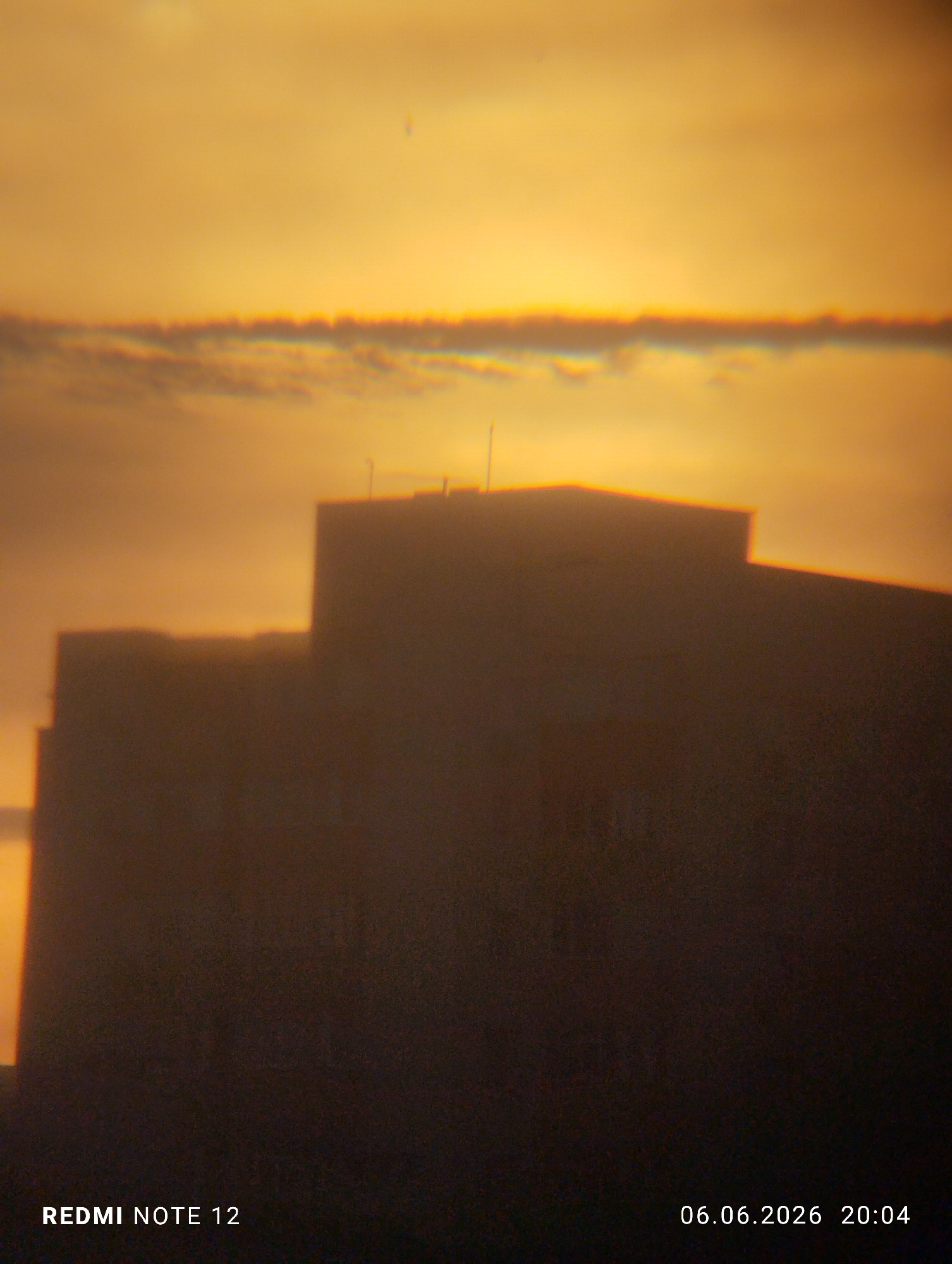 The image depicts an urban landscape at dusk, characterized by a heavily golden-orange sky, likely a sunset, silhouetting foreground structures. The upper portion of the frame is dominated by a bright, warm-toned sky with indistinct cloud formations. A prominent horizontal cloud band in the mid-upper section exhibits significant chromatic aberration, appearing with distinct red and blue fringing along its edges. A faint, small vertical streak is present in the upper-right sky.
In the lower half, two large, dark structures, presumably buildings, are rendered in silhouette against the luminous sky. The building on the right is taller and more defined, presenting a rectangular mass with a flat roofline. Minor protuberances, possibly antennae or small rooftop elements, are visible on its summit. A shorter, less distinct building stands to its left, partially obscured. Both buildings' upper edges, where they meet the bright sky, also show signs of chromatic aberration. No individuals, specific actions, or direct interactions are discernible within the frame.
The image contains textual overlays: "REDMI NOTE 12" in the bottom left corner, indicating the capture device, and "06.06.2026 20:04" in the bottom right, denoting the date and time of capture as June 6, 2026, at 8:04 PM. The specific location within the city of Burgas, Bulgaria, cannot be determined from the visual information presented by the generic building silhouettes.