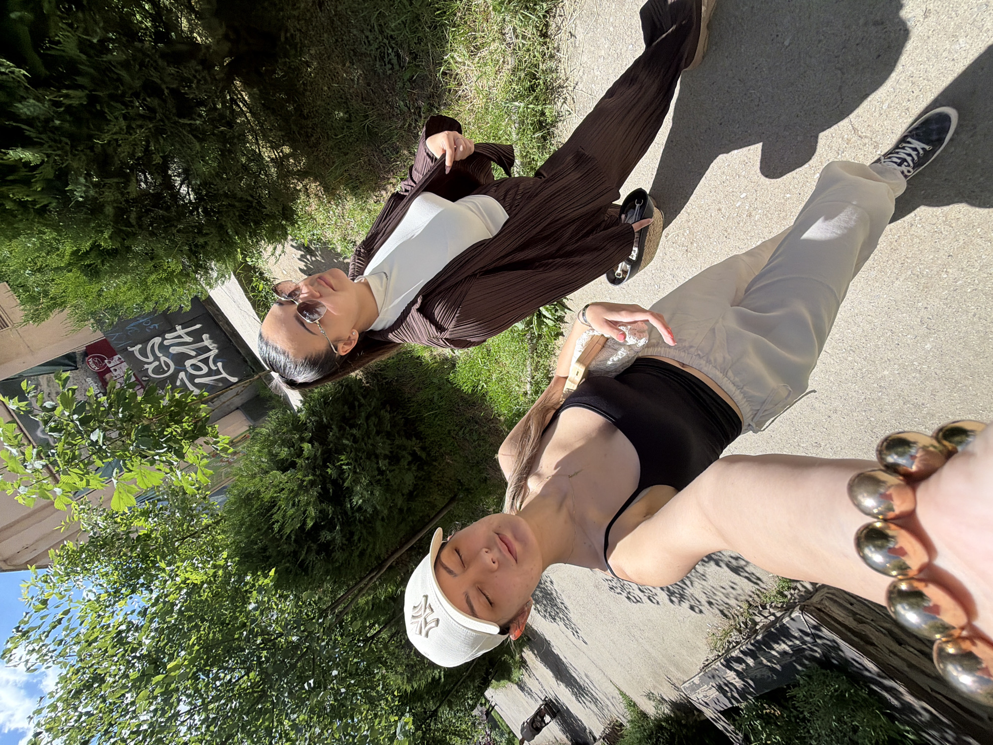 Two women are posing in an outdoor selfie. One wears a white tank top, a brown pinstripe suit, sunglasses, and black shoes. The other wears a black top, beige pants, and white shoes with black laces. She wears a white baseball cap with an 'NY' logo, a gold bracelet, and holds a transparent object in her hand. They are standing on a concrete path near a building, trees, and grass. The location may be in Sofia, Bulgaria.