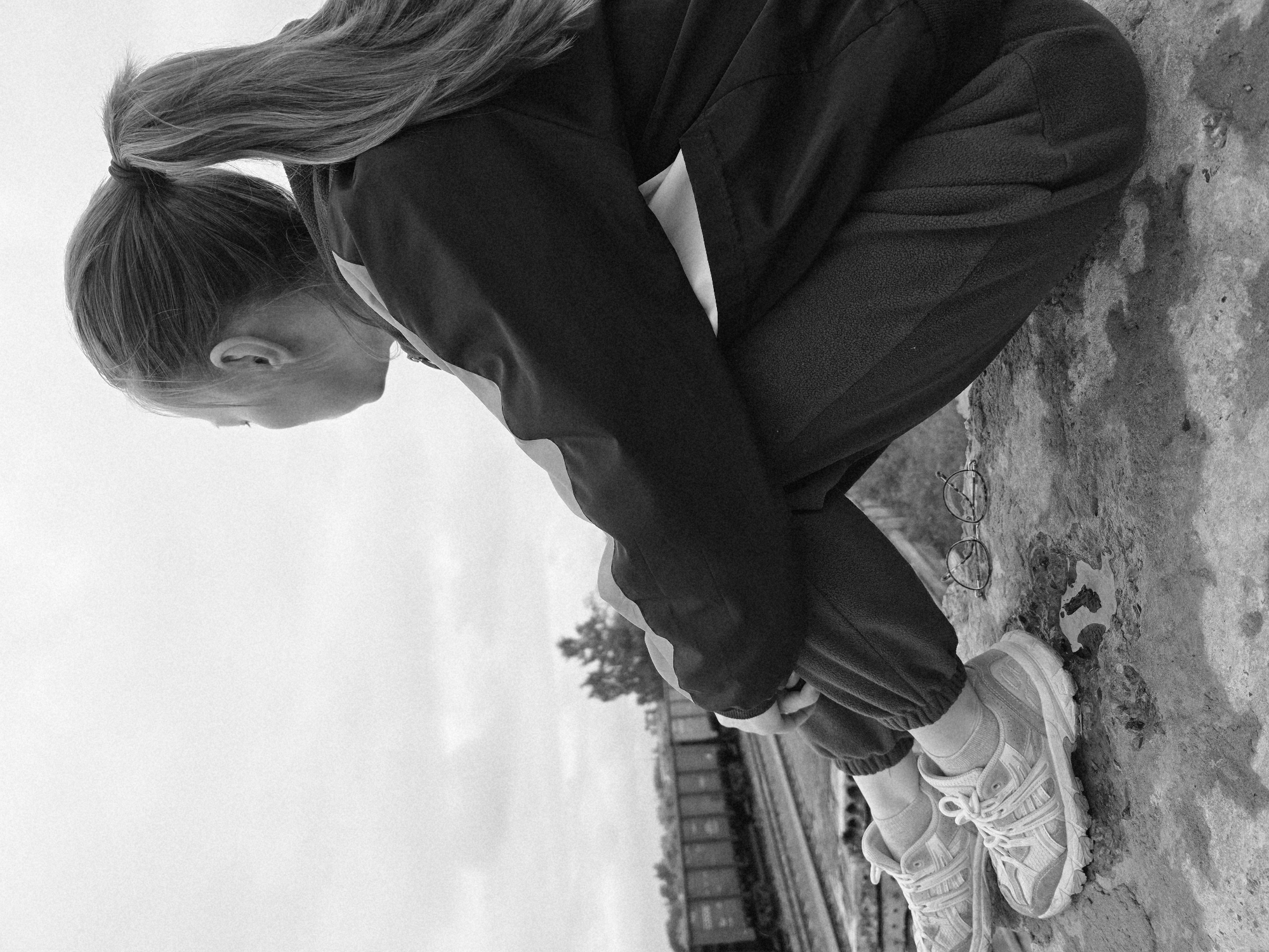 A black and white photograph, captured in Chaikovsky, Russia, depicts a young individual seated in a side-rear profile on a concrete ledge. The person, with light-colored hair pulled into a high ponytail, is dressed in a dark track jacket with lighter stripes on the sleeves, dark sweatpants, and light athletic shoes. Their knees are drawn up, with arms crossed over them, as they gaze towards the right, away from the camera. In the foreground, on the concrete surface near the person's right foot, a pair of wire-frame eyeglasses rests next to a small, irregular puddle of water. The background reveals multiple railway tracks, on which a freight train composed of several boxcars is stationary in the mid-distance. The sky above is overcast, and scattered trees are visible beyond the railway lines, indicating an industrial or transit zone.