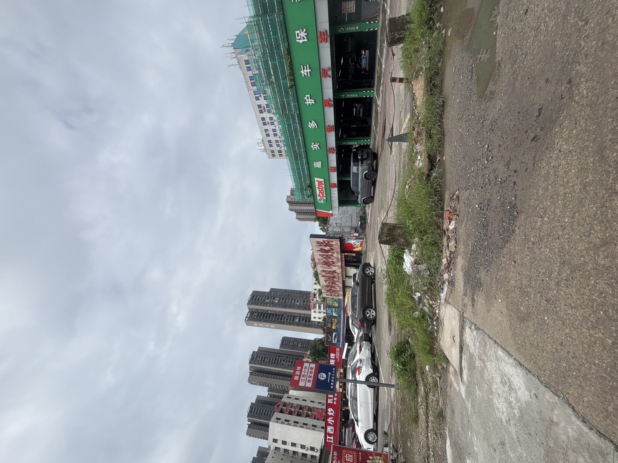 This image shows a street scene in Nanhai District, China, under an overcast sky. In the foreground, a dirt and gravel lot with some patchy grass is visible, littered with trash. A dark gray SUV is parked in front of a green and white building that appears to be a car service center, with "Castrol" branding and Chinese characters indicating repair and maintenance services. Several cars are parked in the bays of the service center, and a construction scaffold is visible on the roof of the building. In the mid-ground, there are several parked cars on a paved area. Behind the service center, a row of tall apartment buildings stretches across the background. Signs with Chinese characters are posted on the buildings, advertising businesses such as restaurants. The overall scene suggests a busy urban environment with commercial and residential areas.