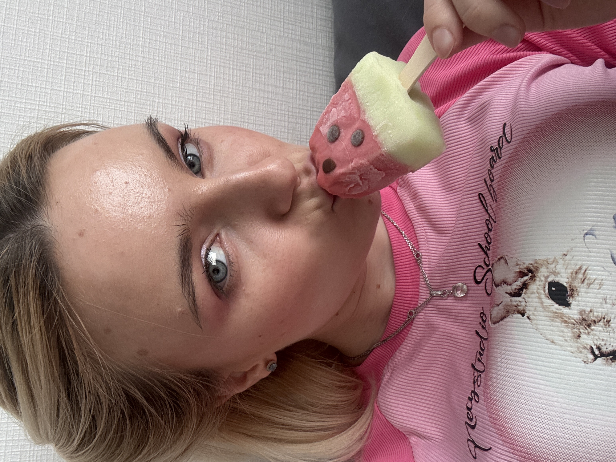 A young woman with light blonde hair and striking blue eyes is depicted from a top-down angle, appearing to lie down or recline. She holds a colorful, character-shaped popsicle near her lips, seemingly about to take a bite or playfully posing with it. The popsicle resembles a watermelon slice or a cartoon character, featuring pink and light green sections, with small dark "eyes" and a "nose" on the pink portion, possibly chocolate chips. It is held by a wooden stick.
She wears a pink, ribbed long-sleeved shirt that transitions to a lighter, white graphic print on the chest area. Visible text on her shirt reads "Precy Studio School Board", alongside a partial image of a light-colored animal's head, possibly a deer or lamb. She has light makeup, including shimmering silver eyeshadow and dark mascara, and wears a silver chain necklace with a clear pendant, as well as a small stud earring. Her expression is focused and slightly playful towards the popsicle, with her lips puckered. The background consists of a plain, light-colored textured wall, indicating an indoor setting.