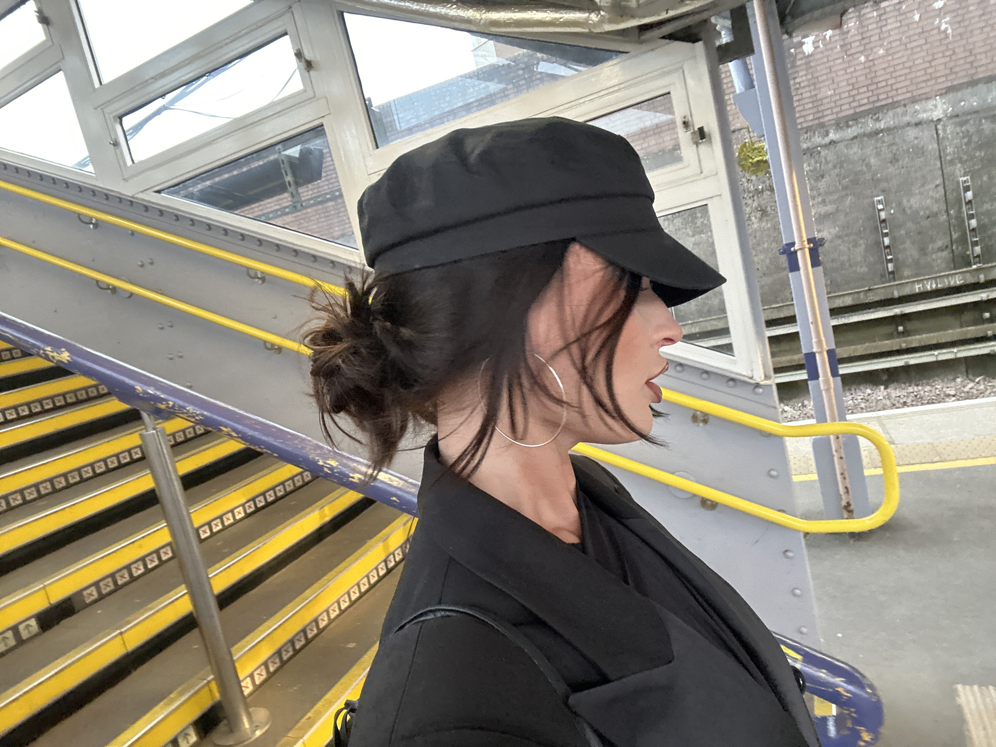 A forensic description of the scene at Tunbridge Wells railway station, United Kingdom, depicts a single individual, likely female, in profile, looking towards the right side of the frame. The individual wears a black captain-style hat, a black blazer over a dark top, and large silver hoop earrings. Dark lipstick is visible on the lips, which are slightly parted. Dark hair is tied back in a bun, with loose strands framing the face.
The individual stands on a grey platform or landing. To the left, a staircase descends away from the viewer. The stairs feature black treads with prominent bright yellow anti-slip strips and small square symbols at the back of each step. Two handrails run parallel to the stairs: a dark purple-blue one in the foreground and a bright yellow one further back. The platform edge and part of the staircase structure are composed of riveted grey metal.
Above the staircase, a multi-pane window structure with white frames provides a view to the exterior. Through these windows, sections of a light brown brick building and a textured grey concrete wall are visible. In the background, beyond the platform, a grey wall section shows the text "HVLIVE" near its base, with horizontal metal strips above it. A metal pole, partially striped blue and white at its base, stands vertically, with a small patch of yellow-green foliage behind it. The ground in the far right background appears to be railway ballast.