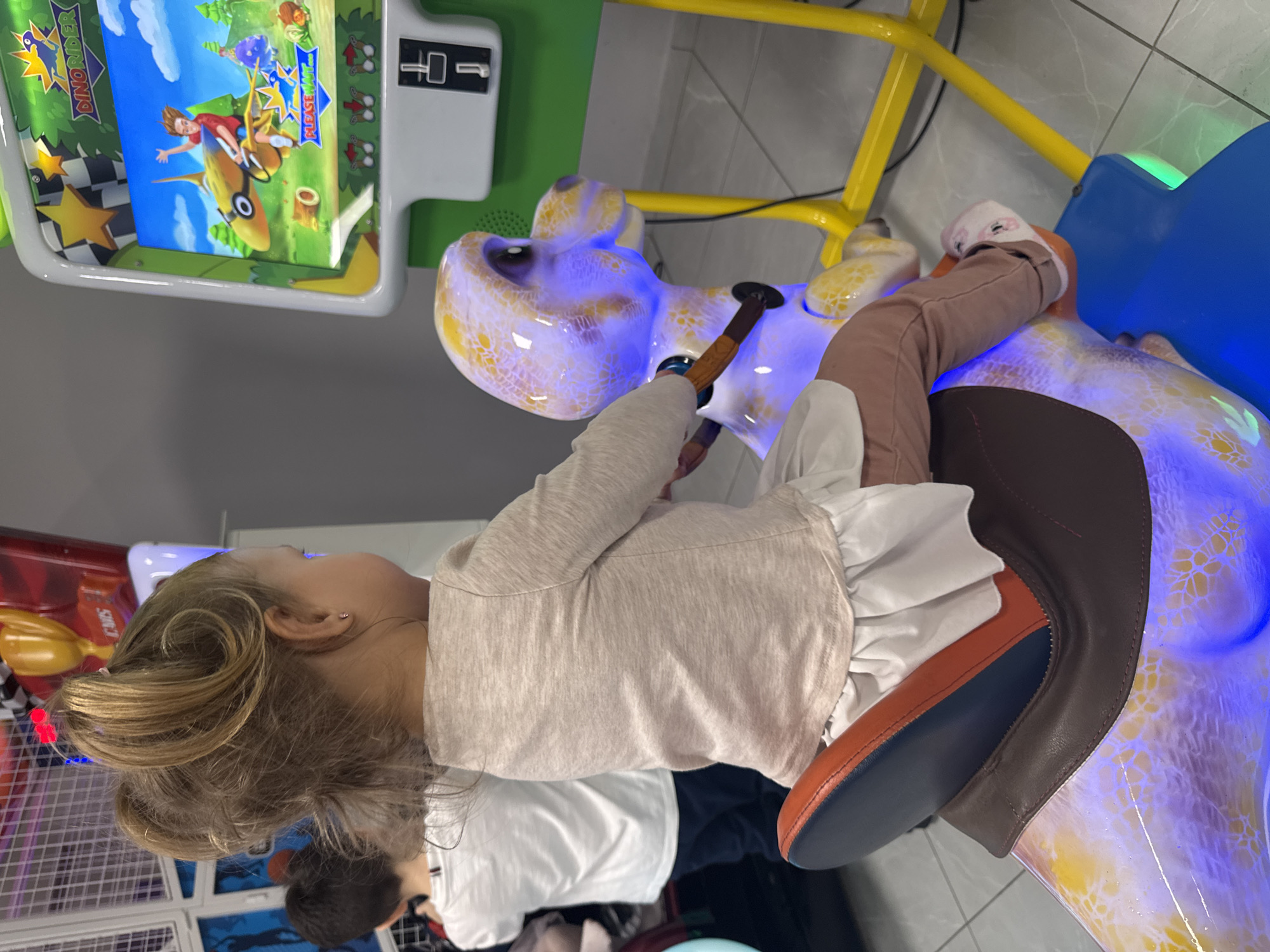 A young child rides a yellow and purple dinosaur coin-operated arcade game in an indoor location, perhaps in the city of Kranevo, Bulgaria. The child is wearing brown pants, a white blouse with ruffles, and a tan sweater. The game has a video screen showing a cartoon character riding a yellow dinosaur, and the game has a yellow metal frame and a blue saddle. A man wearing a white shirt and dark pants is standing behind the child. The background shows arcade machines and a tiled floor.
