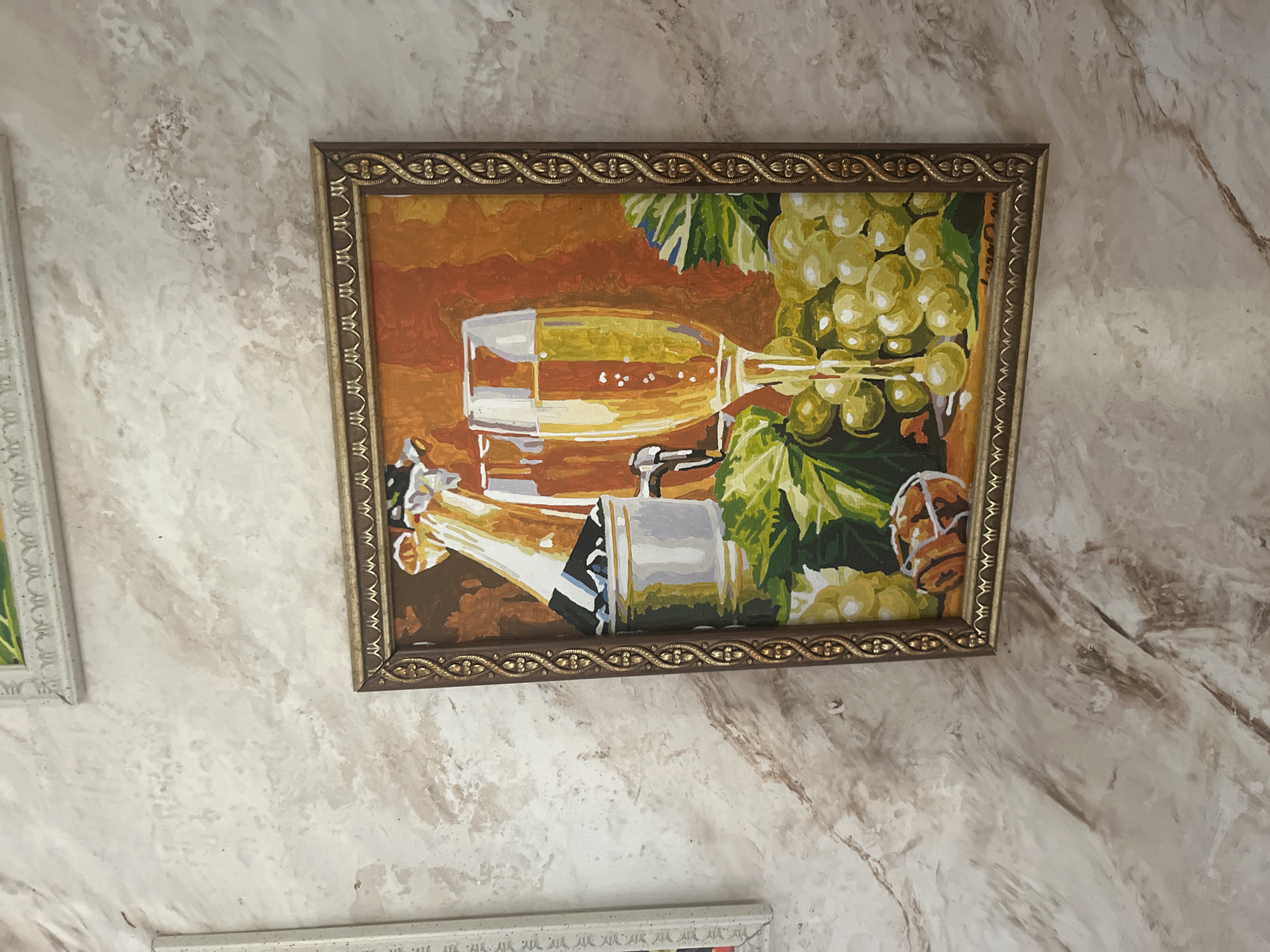 A framed painting of a champagne bottle and glasses with grapes is displayed on a textured wall. The painting is in a dark brown frame with ornate gold detailing. The artwork depicts a close-up view of two champagne flutes, one partially filled with bubbly champagne, and a silver bucket containing a champagne bottle. Clusters of green grapes and leaves are arranged in the foreground. A small, round object, possibly a piece of fruit or a dessert, sits at the bottom of the painting. The wall itself has a marble-like texture with swirling patterns of beige and brown. Another framed artwork with a white border and a green and yellow matting is visible above the champagne painting, partially cropped. The scene is likely indoors, possibly in a home or a commercial establishment. The location in Vladimirovka, Russia, is not explicitly visible in the image.