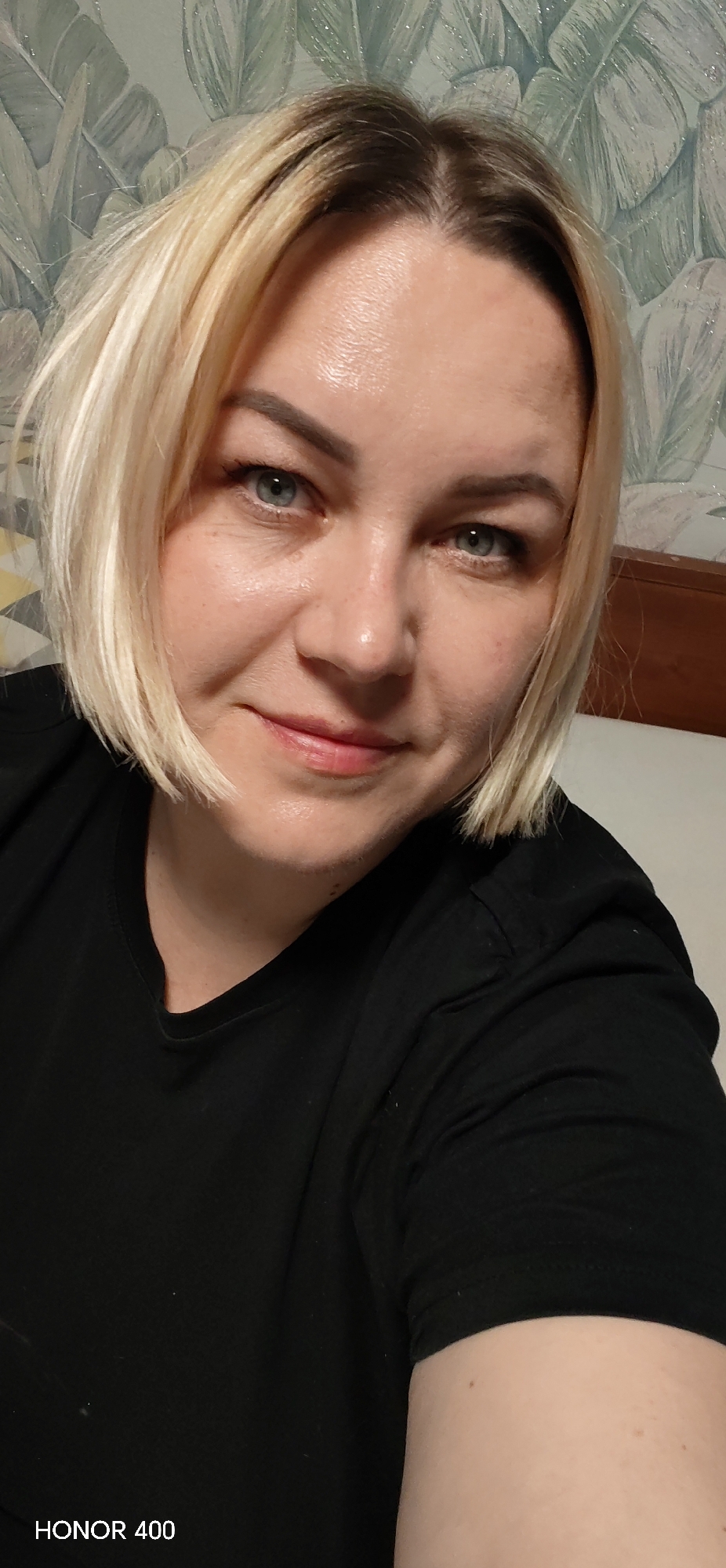 A close-up portrait of a woman with short, blonde hair. She is wearing a black t-shirt. Her eyes are blue. The background consists of a wall with a leaf pattern. The text "HONOR 400" is visible in the lower left corner. The image location is unknown and has not been included.
