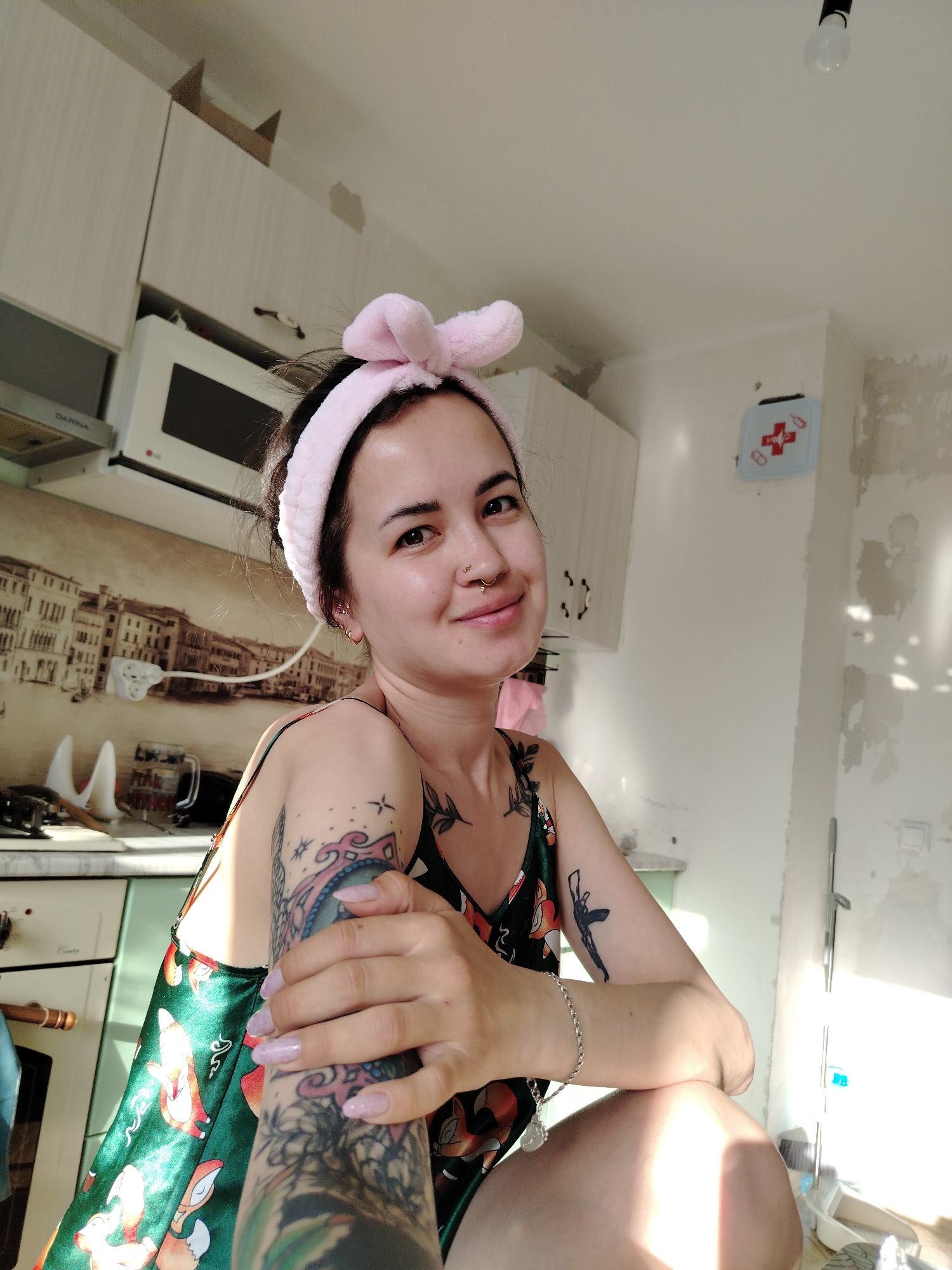 A woman is posing for a selfie in a kitchen. She is wearing a green and red floral print top with a pink bunny ear headband. She has tattoos on both arms and a nose piercing. The kitchen has light-colored cabinets, a microwave, and a stove. The walls are partially plastered and there is a first-aid kit hanging on the wall. The city of Noviy, Russia, is not discernible from this image.