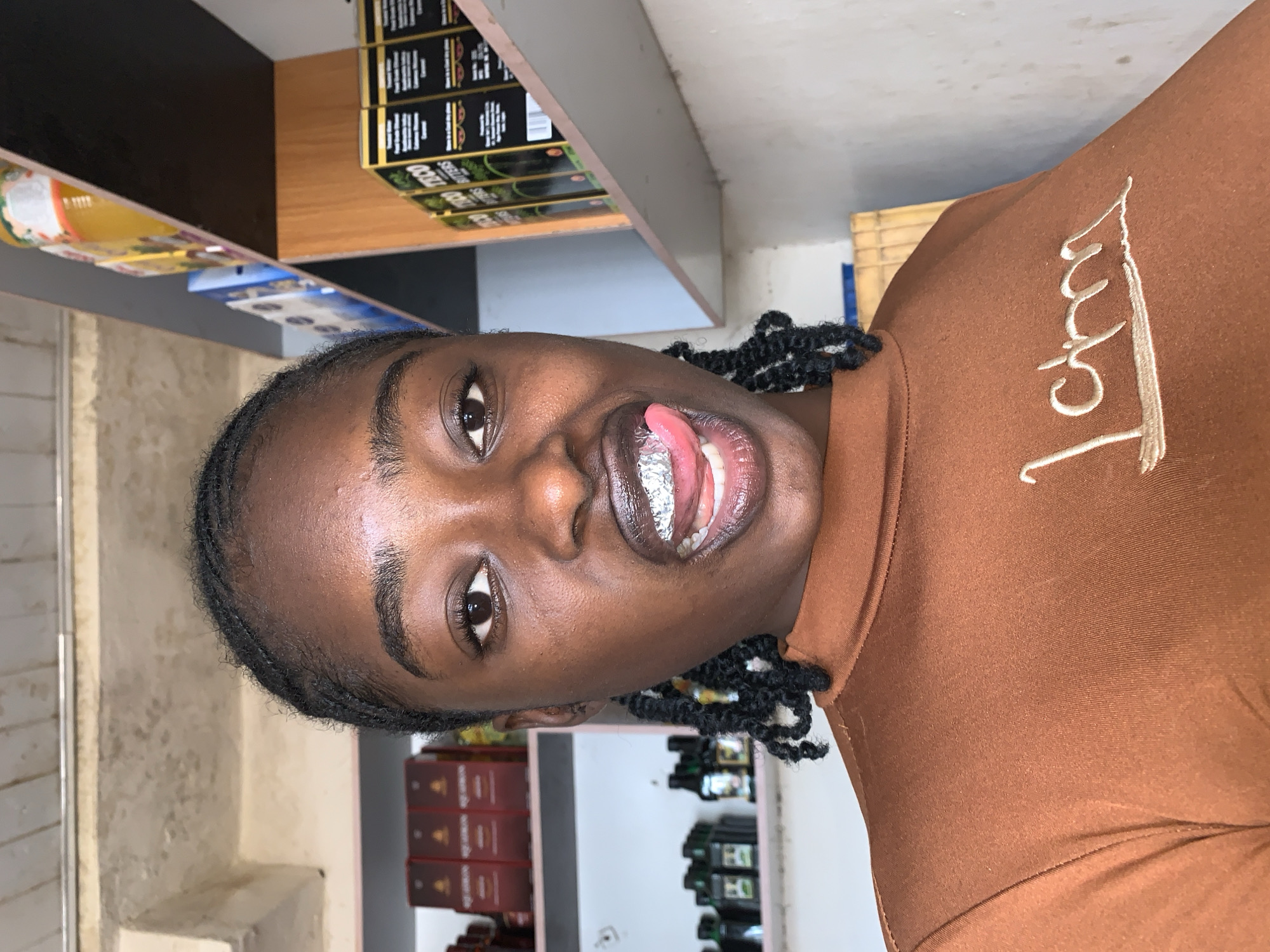 The image captures a head-and-torso shot of a young Black woman, likely in her late teens or early twenties, situated indoors, presumably within a retail establishment, in New Stadium, Nigeria. She has dark skin, dark brown eyes, and dark braided hair pulled back from her face. Her gaze is directed straight at the camera. Her mouth is open, with her tongue extended and pressing against a reflective, crumpled, metallic-appearing object that covers her upper front teeth. She wears a fitted, long-sleeved top in a burnt orange or terracotta color with a high neckline. Embroidered on the chest of her top, in a lighter tone, are the words "I am" in a script font, underlined.
The background consists of shelving units on both sides of the subject. To her left, an upper shelf displays multiple dark red-brown rectangular boxes, while a lower shelf holds rows of small, dark-colored bottles. To her right, an upper shelf contains various packaged goods, including a yellow-orange container and items in blue packaging. A middle shelf, positioned around the subject's eye level, features several black rectangular boxes with green labels, where the text "TECO BUTTERS" is visible. The walls are light-colored and exhibit a textured, possibly concrete, surface. A portion of the ceiling, showing exposed concrete or wooden beams, is visible in the upper left corner. The scene is illuminated by ambient light.