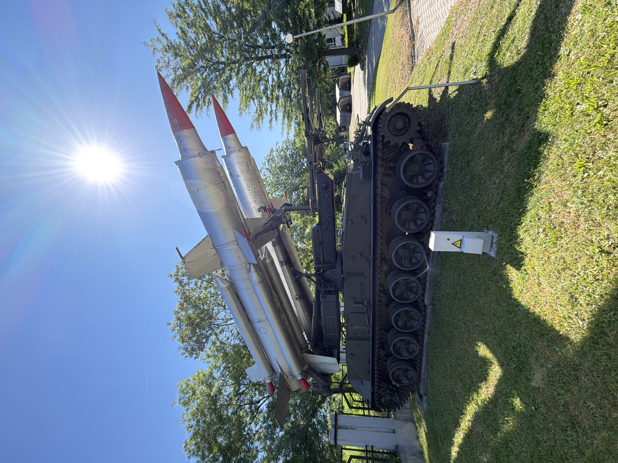A surface-to-air missile launcher, mounted on a tank chassis, is displayed outdoors under a clear blue sky with a bright sun. The military vehicle, painted olive drab, is situated on a grassy hill, with the missile launcher loaded with two long, silver missiles tipped with red. The missiles are arranged diagonally, one above the other. In the background, several mature trees with lush green foliage are visible, along with a paved walkway and some buildings. A tall lamppost stands to the right of the vehicle. A small electrical box with a yellow triangle warning symbol is present in the foreground. The location is the Museum of Socialist Art in Sofia, Bulgaria.