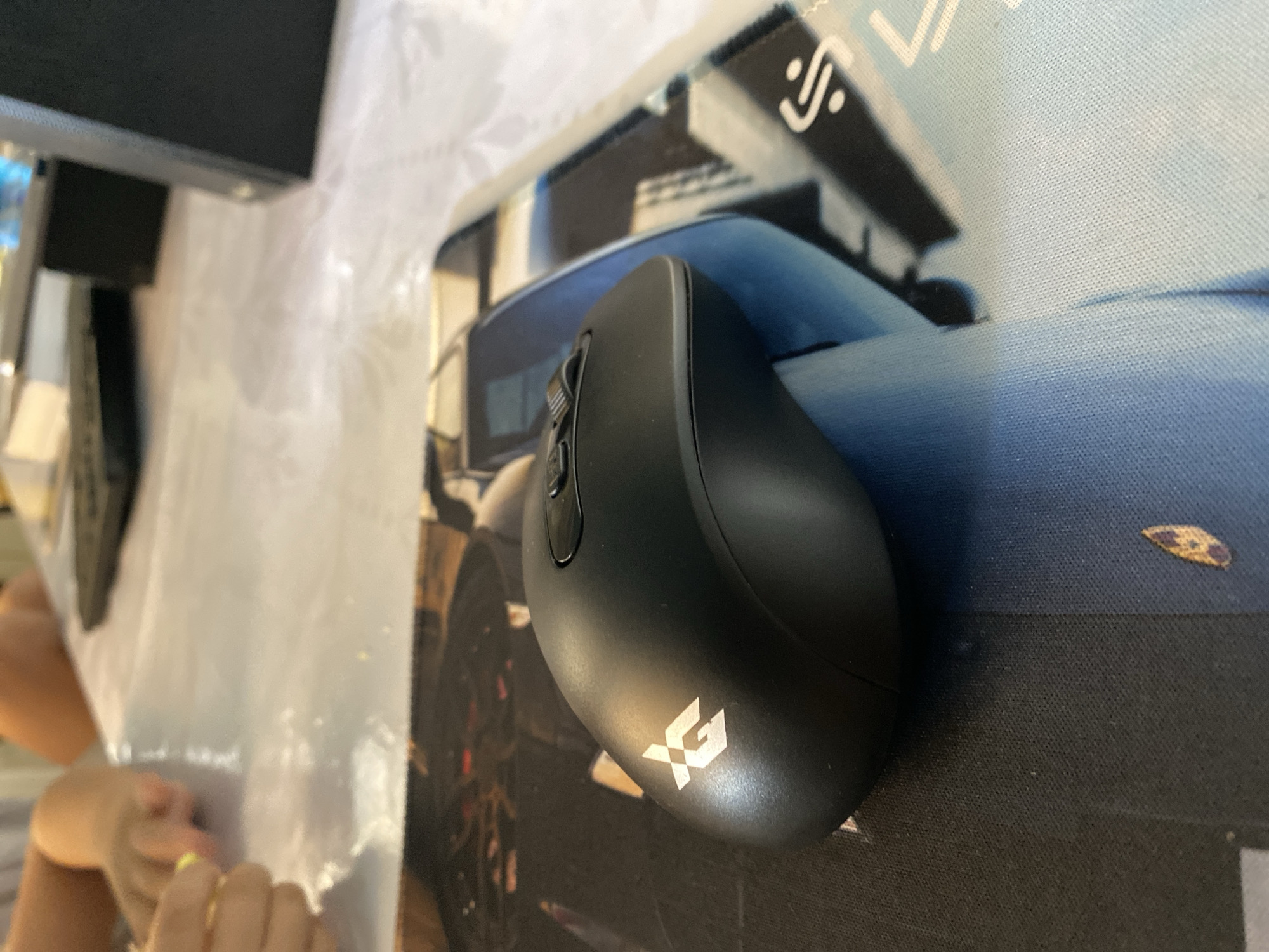 An overhead close-up shot shows a black computer mouse with the white logo "GX" on its surface. The mouse is resting on a mousepad with a blue and black abstract design, and the mousepad is on a table covered with a white plastic tablecloth. In the background, blurred figures of people's arms are visible, along with a laptop and a black object that appears to be a computer tower. The scene is indoors.