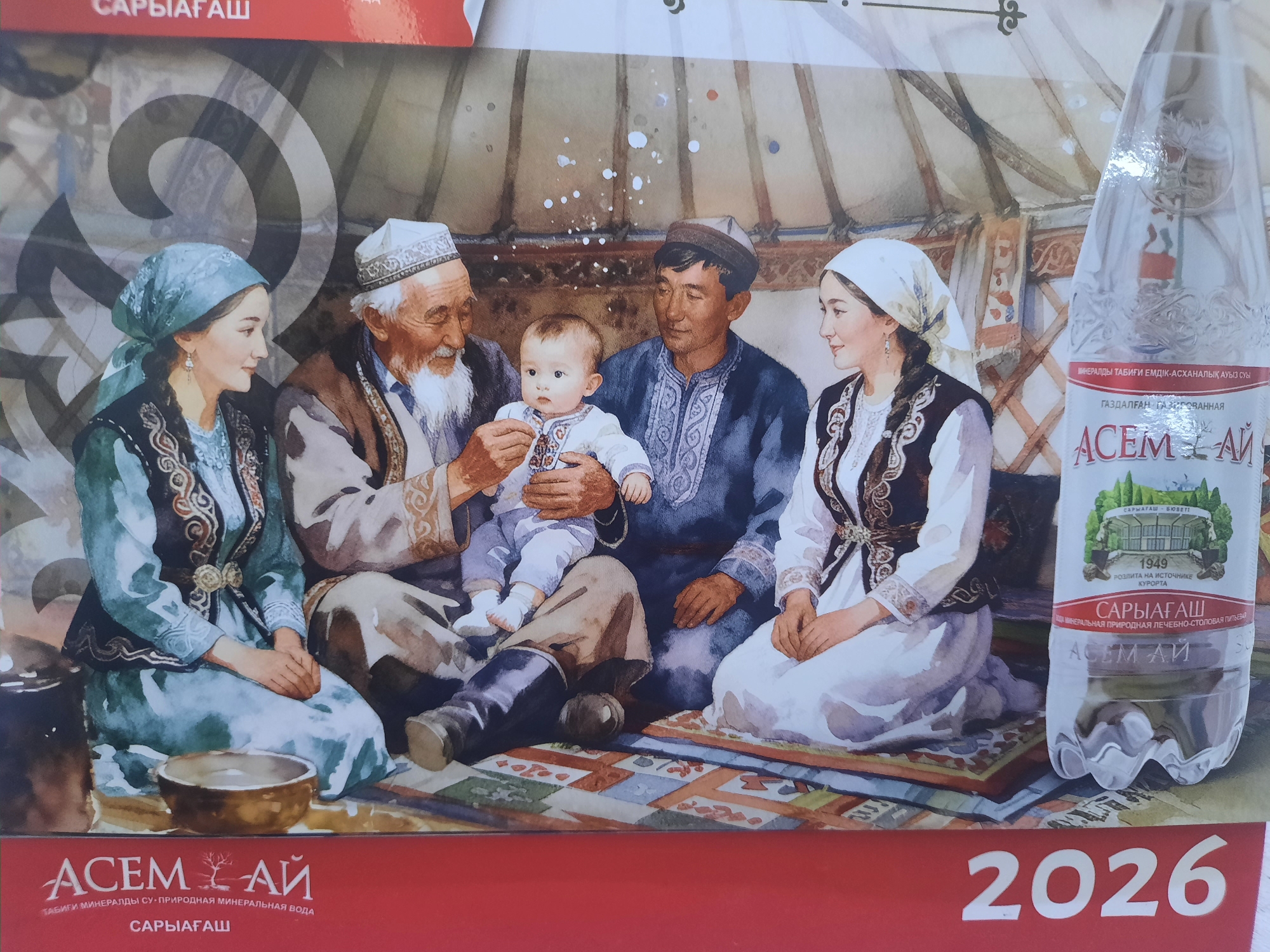 This is a digital image depicting a watercolor painting of a family in traditional Kazakh clothing gathered inside a yurt. On the right side of the painting, a bottle of "Асем-Ай" mineral water is prominently displayed. The painting shows an elderly man with a white beard, a man in a blue garment, a woman in a green dress, and another woman in a white headscarf and black vest, all looking towards a baby being held by the elderly man. The background features the interior of a yurt with its characteristic lattice walls and wooden poles. The lower portion of the image displays the brand name "Асем-Ай" and the text "САРЫАГАШ" (Saryagash), which refers to a resort town in Turkestan Region, Kazakhstan, known for its mineral water. The year "2026" is visible in large white numerals on a red background at the bottom right of the image. While the prompt mentions the city of Zhanaozen, Kazakhstan, this specific image does not contain any visual elements that definitively place the scene within Zhanaozen. The primary identifiable location indicated by the text is Saryagash, Kazakhstan, known for its mineral water brand "Асем-Ай".