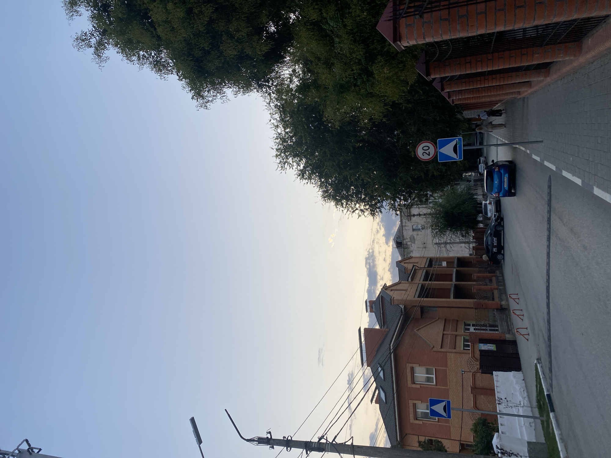 The image depicts a street scene in Feodosia, Ukraine. A two-story brick house with a dark roof is visible on the left side of the street. A street lamp with multiple wires extends from a pole on the left. Further down the street, a blue car is parked on the right side of the road, next to a sidewalk. Traffic signs, including a speed limit sign for 20 km/h and a sign indicating a speed bump, are posted along the sidewalk. Two individuals are walking on the sidewalk. A brick fence lines the right side of the street. The sky is a pale blue with some clouds, suggesting it is either early morning or late afternoon. The overall environment appears to be a residential area.