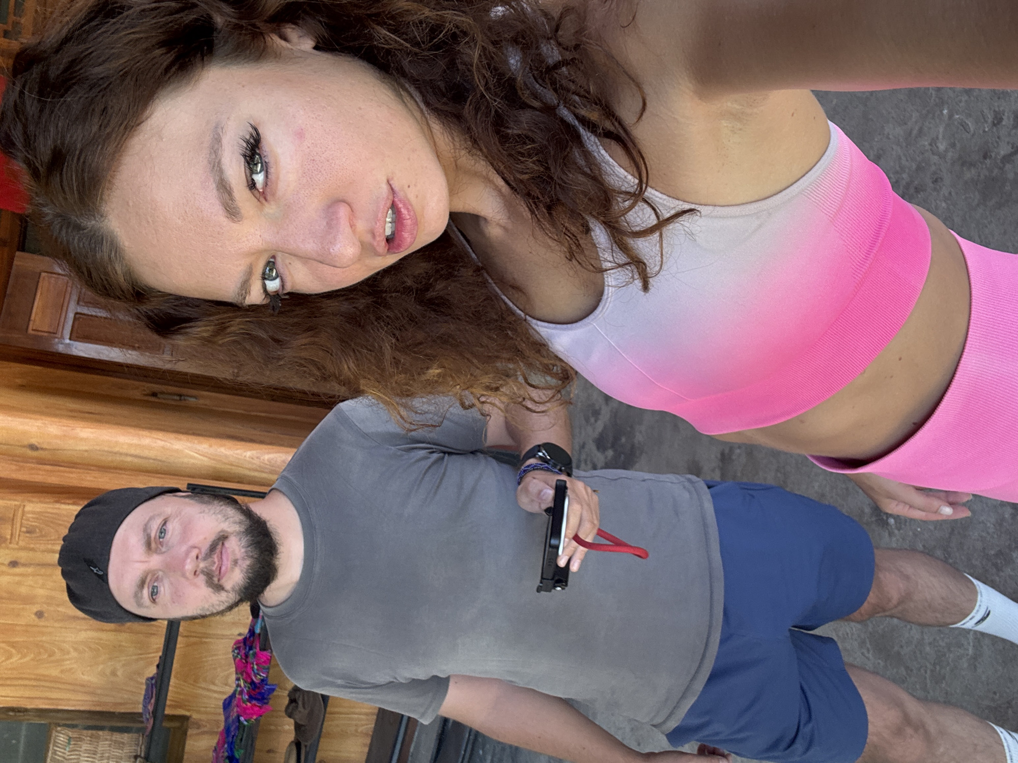 A selfie photograph shows a Caucasian woman and man. The woman has curly brown hair, fair skin, and green eyes. She wears a pink-and-white ombre athletic top. The man has a beard, blue eyes, and wears a black beanie, gray t-shirt, and blue shorts. He holds a small, black object with a red string. They stand outdoors in Bắc Hà, Vietnam, evidenced by the local architecture and the presence of regional textiles.