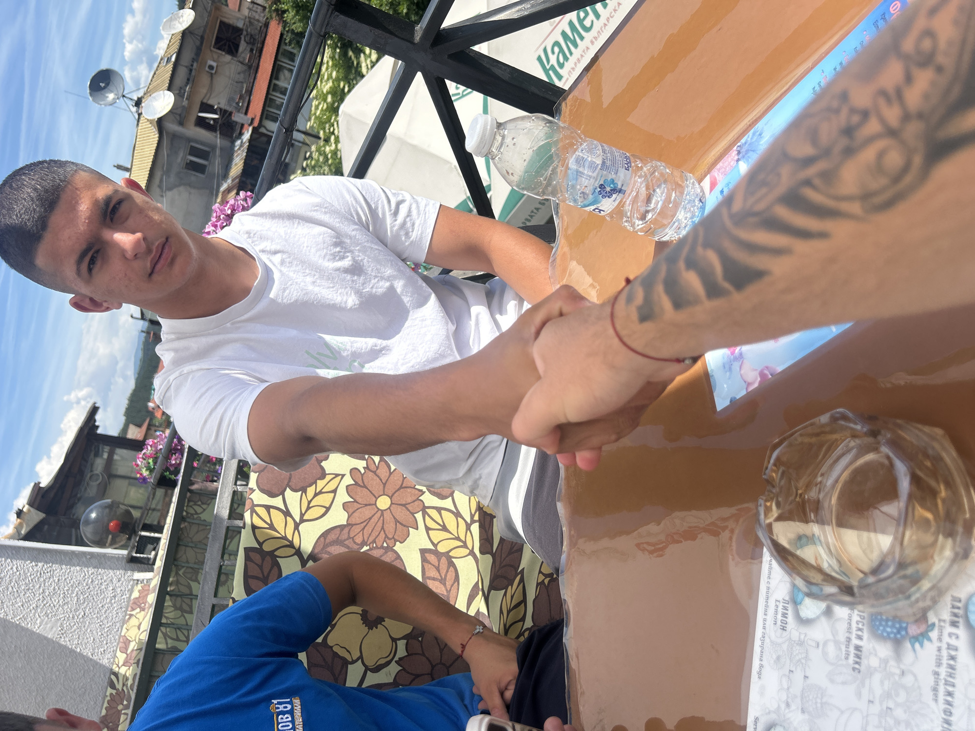 An outdoor scene in Bania, Bulgaria, shows two men shaking hands across a shiny, light brown table. The man on the left, with short dark hair and wearing a white t-shirt, looks directly at the camera. He has visible acne on his face. The other man's arm, heavily tattooed with a full sleeve design and adorned with a red string bracelet, extends from the right side of the frame to complete the handshake. A third person, wearing a blue t-shirt with a white logo, is partially visible at the bottom left, seated on a floral-patterned bench.
On the table rests a plastic water bottle labeled "БАНКЯ" (Bankya) and "500ml", along with an empty glass ashtray. Beneath the ashtray is a menu or paper displaying drink options such as "ЛАЙМ С ДЖИНДЖИФИЛ" (Lime with ginger), "ОРСКИ МИКС" (Forest fruits), and "Лимон" (Lemon), with a note about serving with still or sparkling water.
The background reveals a sunny day with a clear blue sky and some clouds. Behind the man in white, a building with multiple satellite dishes on its roof and vibrant purple flowers in window boxes is visible. Further in the background, a large white umbrella with "Камена" (Kamena) and "ПЪРВАТА БЪЛГАРСКА" (The first Bulgarian) branding is partly obscured, suggesting a cafe or restaurant setting. The strong lighting and shadows indicate it is likely midday or early afternoon. The atmosphere appears casual.