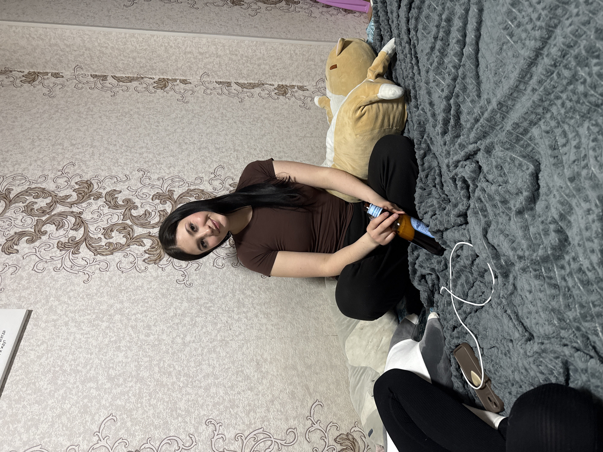 A woman is sitting on a bed against a wall with patterned wallpaper. She is wearing a brown shirt and black pants and is holding a bottle. A stuffed animal sits next to her against a gray textured blanket that covers part of the bed. An off-white object is visible to the left. A cell phone on the bed.