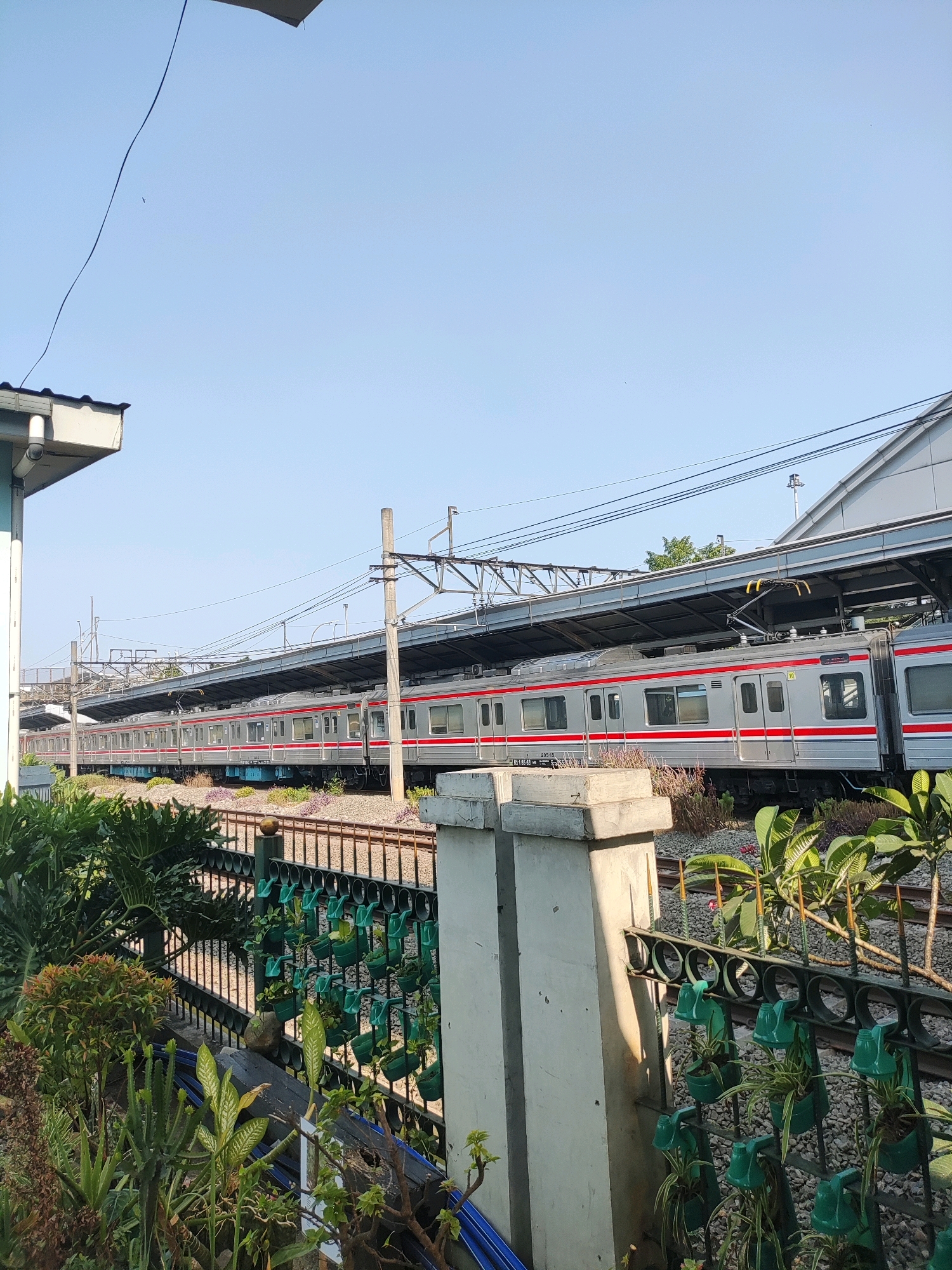 The image shows a train at a station platform on a clear day. The train is silver with red stripes and has multiple carriages. The platform is covered by a grey roof supported by pillars. There are overhead wires and electrical equipment associated with the train line. In the foreground, there is a black metal fence with small green planters hanging from it, containing various plants. Behind the fence, there are railway tracks and ballast. Lush green foliage and some concrete structures are also visible in the foreground. The background is a clear blue sky. This appears to be a scene from the city of East Jakarta, Indonesia, likely at a train station. No people are visible in the image.