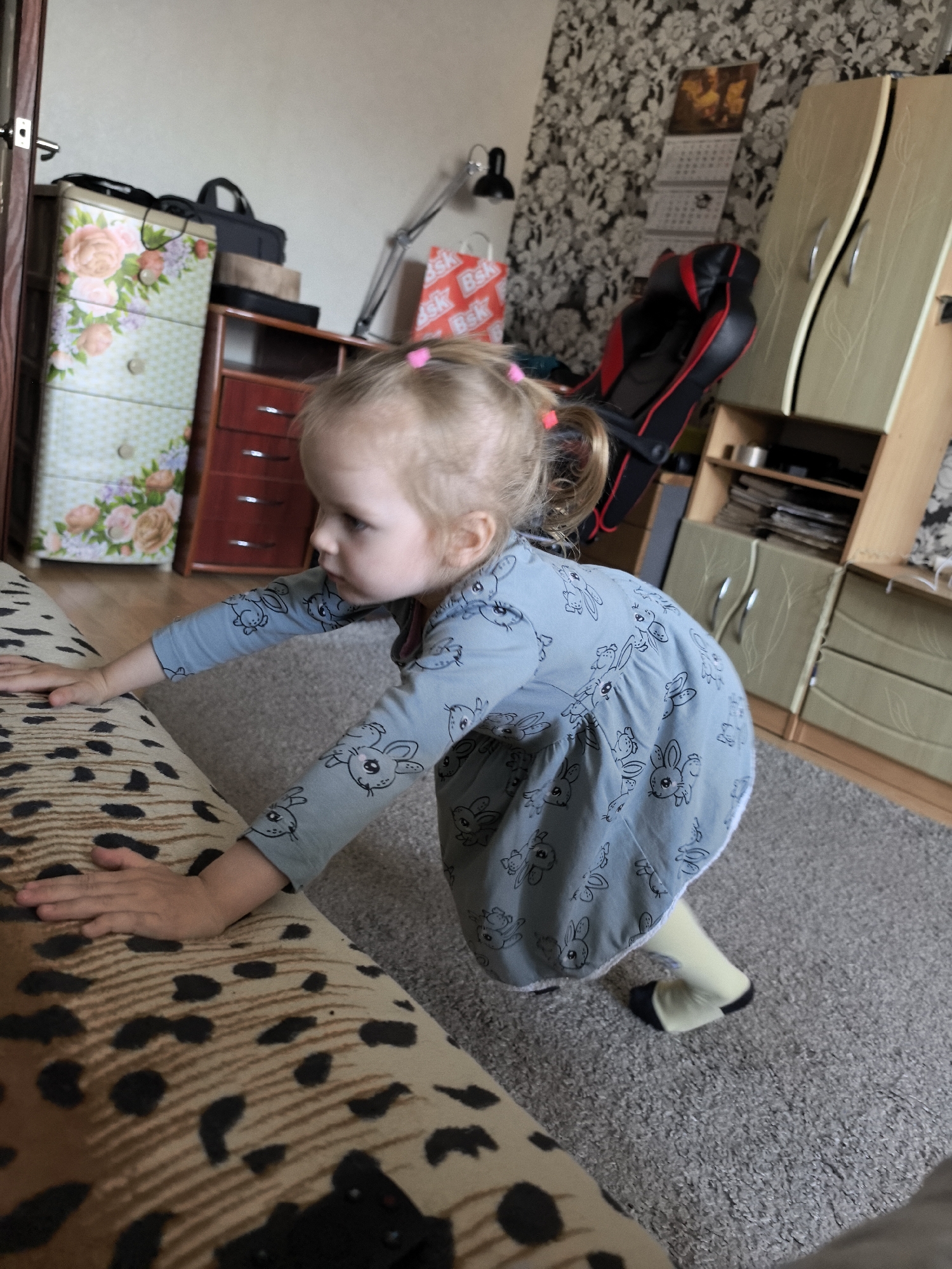A young girl with blonde pigtails is on her hands and knees, pushing off of a sofa. She is wearing a light blue, long-sleeved onesie with a pattern of black rabbits. Her legs are clad in light yellow tights and black-tipped socks. The sofa has a spotted pattern in brown and black. The background of the image shows an indoor scene, possibly a child's bedroom or play area. To the left, a chest of drawers is decorated with floral patterns. To the right, a wardrobe with cream-colored doors stands against a wallpapered wall. A black and red gaming chair is visible near the wardrobe, and a desk lamp is positioned behind the girl. A calendar hangs on the wall. The lighting suggests it is daytime.