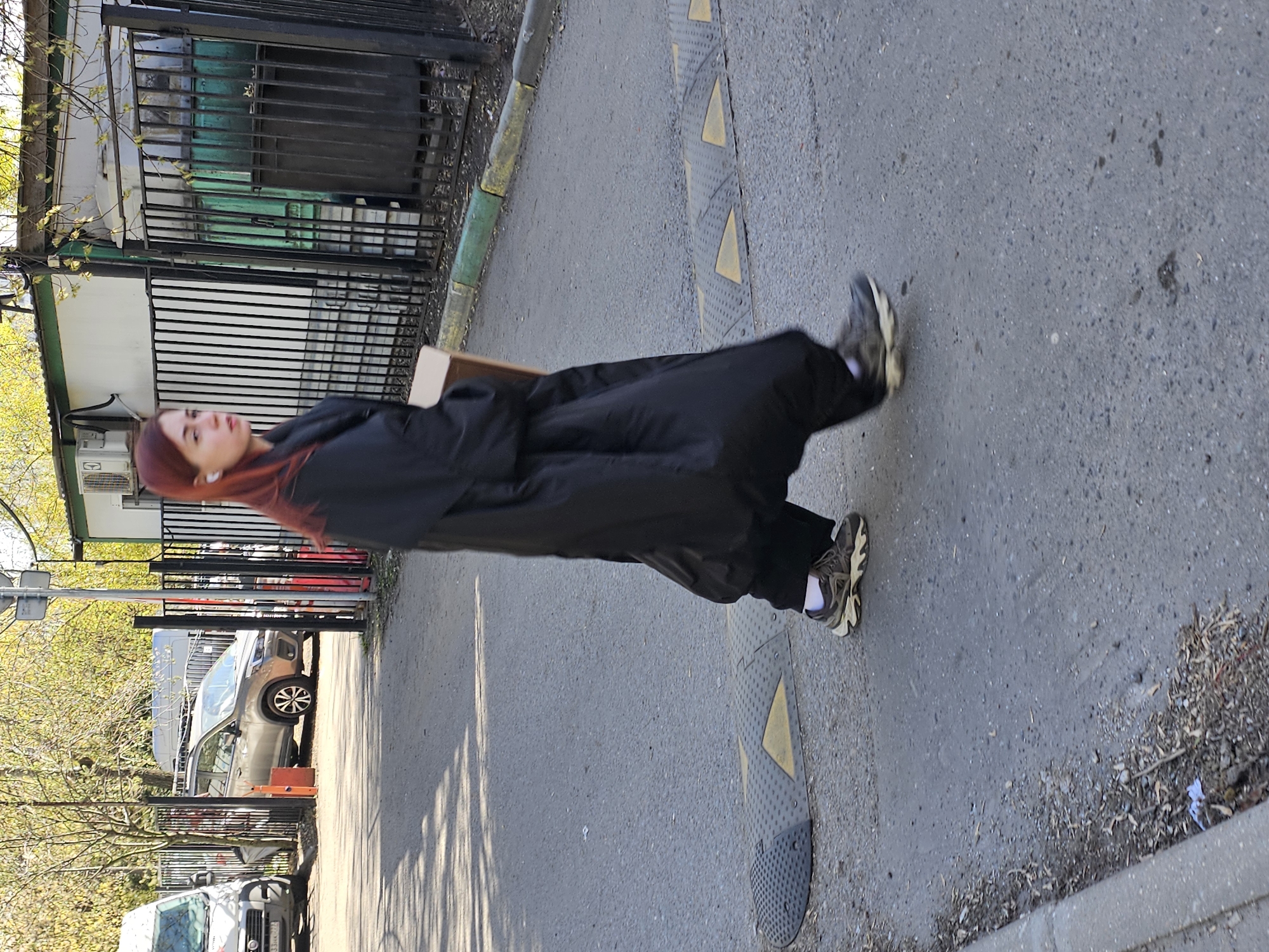 A young woman with auburn hair, wearing a black robe or long coat and casual athletic shoes with white socks, walks on a gray concrete sidewalk. Speed bumps are visible on the side of the sidewalk with a yellow-and-gray pattern. The scene appears to be in an urban environment, likely Moscow, Russia. A fence is situated nearby, with cars parked beyond it. Trees are present with yellowing leaves, suggesting it might be autumn. The woman seems to be walking at a normal pace on a sunny day.