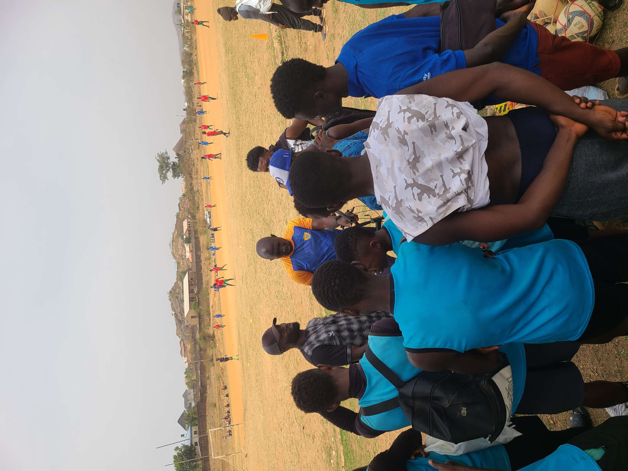 The image shows a group of people in an open field, likely in Barikin Jirga Summa, Nigeria. They are standing in a huddle, possibly observing a soccer game in progress on the field. Some individuals are wearing soccer jerseys, with players visible on the field in red and blue uniforms. There are soccer balls near the group. In the background are buildings and a rocky hill or outcrop. The sky is overcast.