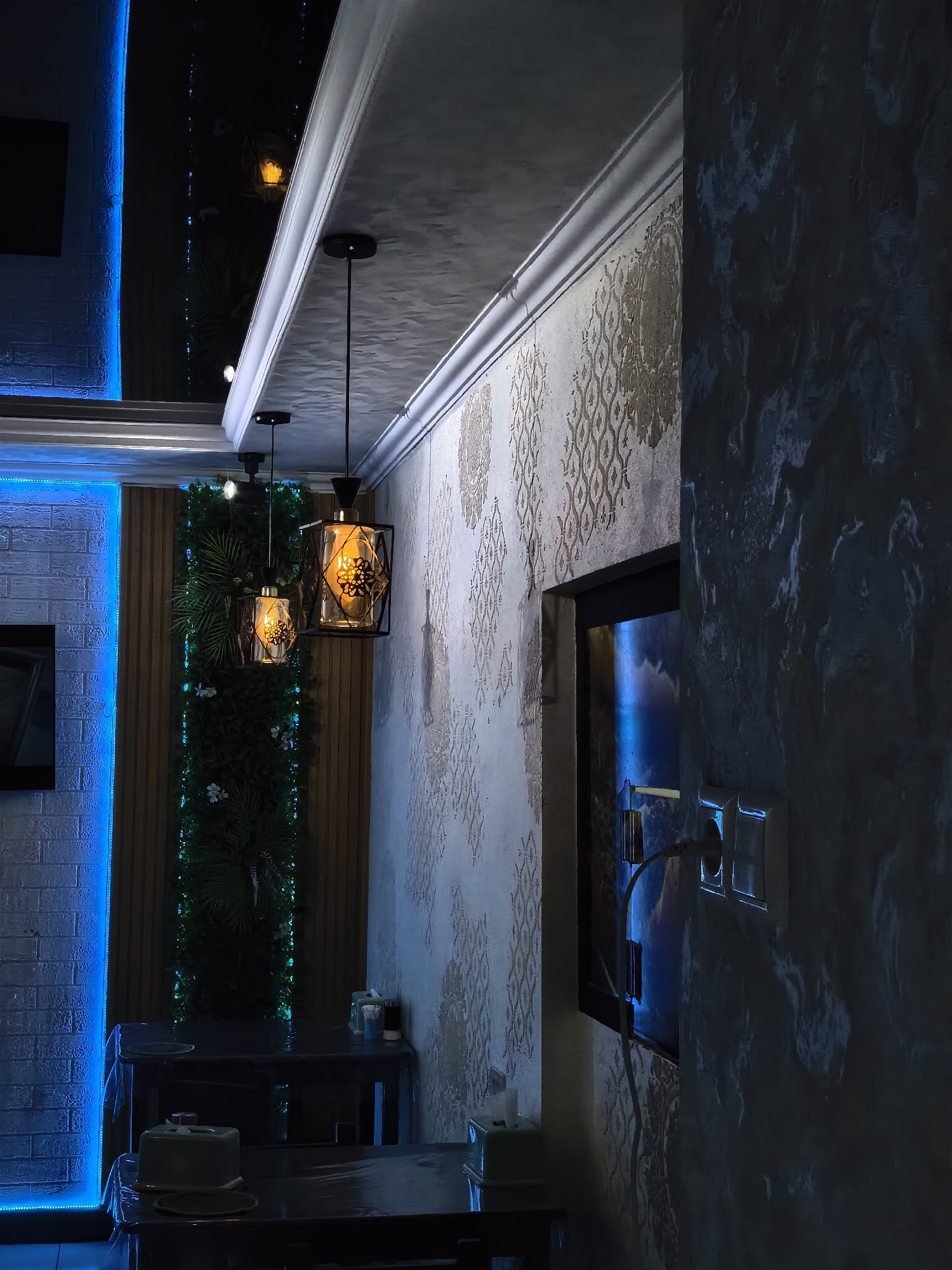 The image depicts an interior scene, likely a restaurant or cafe, with a modern and dimly lit ambiance. The foreground features two dark tables covered with clear plastic sheeting. On the tables, there are tissue box holders and some other unidentifiable objects. In the background, a vertical green wall adorned with artificial plants and small lights is visible. Two pendant lights with black metal frames and amber glass shades hang from the ceiling.
The walls of the establishment are decorated with a textured, light-colored wallpaper featuring a repeating ornate pattern. To the left, a brick wall is illuminated by bright blue LED strip lights. The ceiling is dark and reflective. A framed screen is mounted on the wall, displaying a vibrant, possibly abstract image with blue hues. An electrical outlet with a plug and cable is visible on the wall near the screen. The overall lighting is atmospheric, with a mix of blue, amber, and white light sources contributing to the mood. The scene appears to be devoid of any people. The location is identified as Fergana City, Uzbekistan.