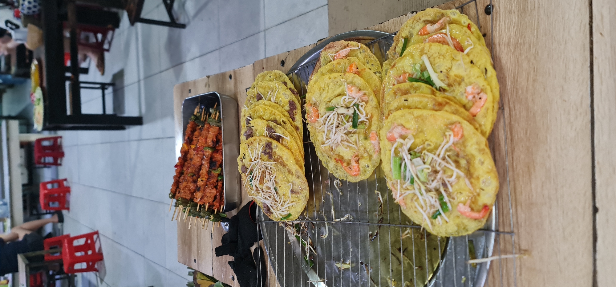 The image displays a vibrant street food stall, likely in Đà Nẵng, Vietnam, showcasing a variety of prepared dishes. The foreground is dominated by two distinct types of food arranged on a wooden counter.
On the right, numerous round, yellow, savory pancakes are displayed on a metal grill rack. These pancakes are generously topped with ingredients: many feature bright pink shrimp, fresh white bean sprouts, and green scallions, characteristic of Vietnamese Bánh Khọt. Other pancakes appear to be topped with a darker, finely chopped ingredient, possibly minced meat. The pancakes are stacked and neatly arranged, ready for consumption.
To the left of the pancakes, a rectangular metal tray holds a quantity of grilled skewers. These skewers are loaded with reddish-orange marinated meat, interspersed with green vegetables or herbs, indicating they are freshly cooked and seasoned. The wooden skewers are visible, suggesting they are single portions.
In the background, the setting appears to be a casual outdoor or semi-outdoor eating area. Several red plastic stools are scattered, along with dark-colored tables or table frames, typical of Vietnamese street eateries. A person's legs and partial body are visible sitting at one of the tables on the far left. The flooring is a light-colored tiled surface.
The scene is well-lit, possibly by artificial lights, common for evening street food vendors. The focus is clearly on the appetizing food presentation, inviting customers. There is no visible text in the image.