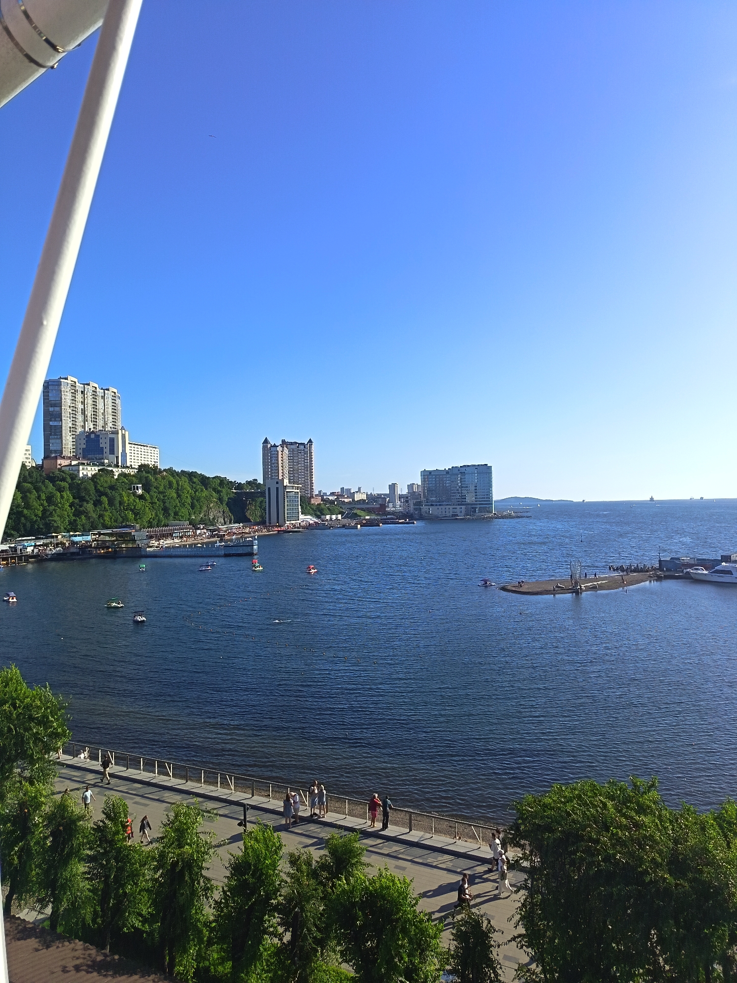 The image shows a bright, sunny day with a clear blue sky overlooking a body of water. In the foreground, a white structure, possibly part of a large building or a ferris wheel, is visible on the left. A paved walkway runs along the waterfront, with people walking and standing on it. Lush green trees line the edge of the walkway, casting shadows on the pavement.
Across the water, a cityscape with several tall buildings is visible on a green, hilly terrain. One prominent building is a tall, modern, multi-story residential or office complex. Further along the waterfront, a pier extends into the water, with more buildings and a docking area. Small boats and paddleboats are scattered across the water. In the distance, more buildings and hills can be seen, and a few ships are visible on the horizon. The overall scene depicts a vibrant urban waterfront with recreational activities and modern architecture.