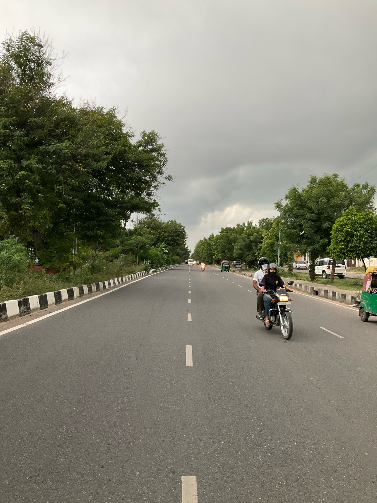 The image shows a road in the Jaipur Municipal Corporation, India, on a cloudy day. The road is paved with asphalt and has white dashed lines marking the lanes. On the left side of the road, there are trees and vegetation. On the right side, there is a sidewalk with a black and white striped border, followed by more trees, parked cars, and buildings in the distance. Two people on a motorcycle are riding in the right lane, facing away from the camera. The person in the back is wearing a black shirt and a white mask. The person in the front is wearing a black t-shirt and a helmet. A green auto-rickshaw is visible in the far right of the image. The sky is overcast and appears to be a dark gray color.