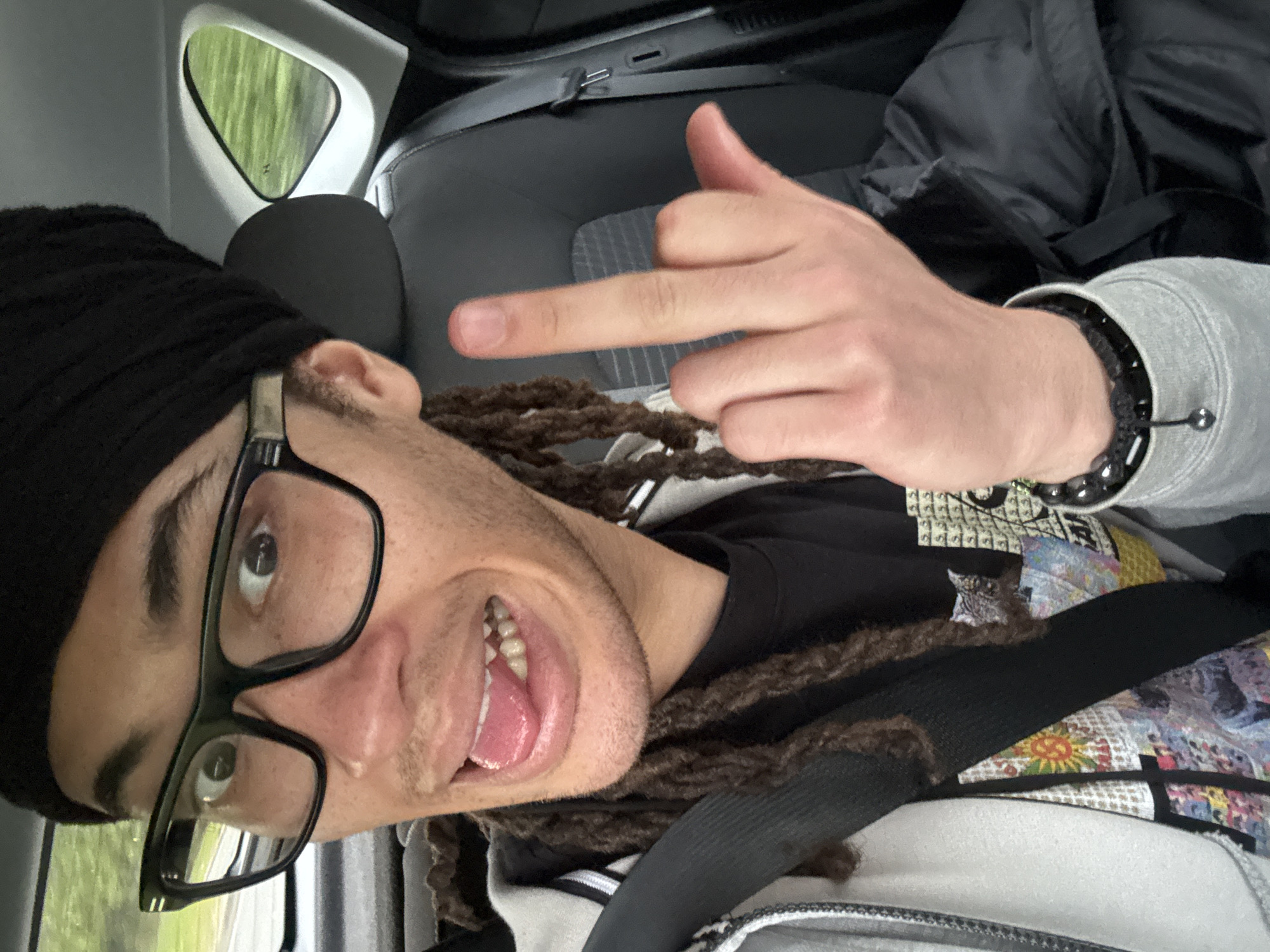 A man is seated in a car, wearing a black beanie, black glasses, a black t-shirt with a graphic design, and a light gray jacket. He is wearing a bracelet on his left wrist and has dreadlocks. He is wearing a seatbelt and is giving the middle finger. The car's interior is visible, including the seats and a small triangular window. The scenery visible through the window suggests a green, outdoor environment. There is no information about the image location.