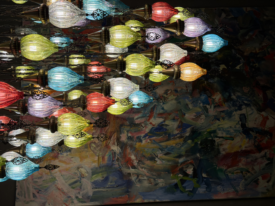 The image displays a multi-tiered chandelier consisting of numerous illuminated glass lanterns. Each lantern features a ribbed, bulbous glass body in various colors including red, orange, yellow, lime green, blue, cyan, purple, and clear, capped with decorative brass-toned metal finials. The lanterns are suspended at varying heights by dark metal chains and ornate filigree elements, creating a dense cluster of light. In the background, to the left and upper-left, a large abstract painting is partially visible, characterized by broad, dynamic brushstrokes in colors such as blue, green, red, white, and yellow. The scene is located indoors, within the city of Budva, Montenegro.