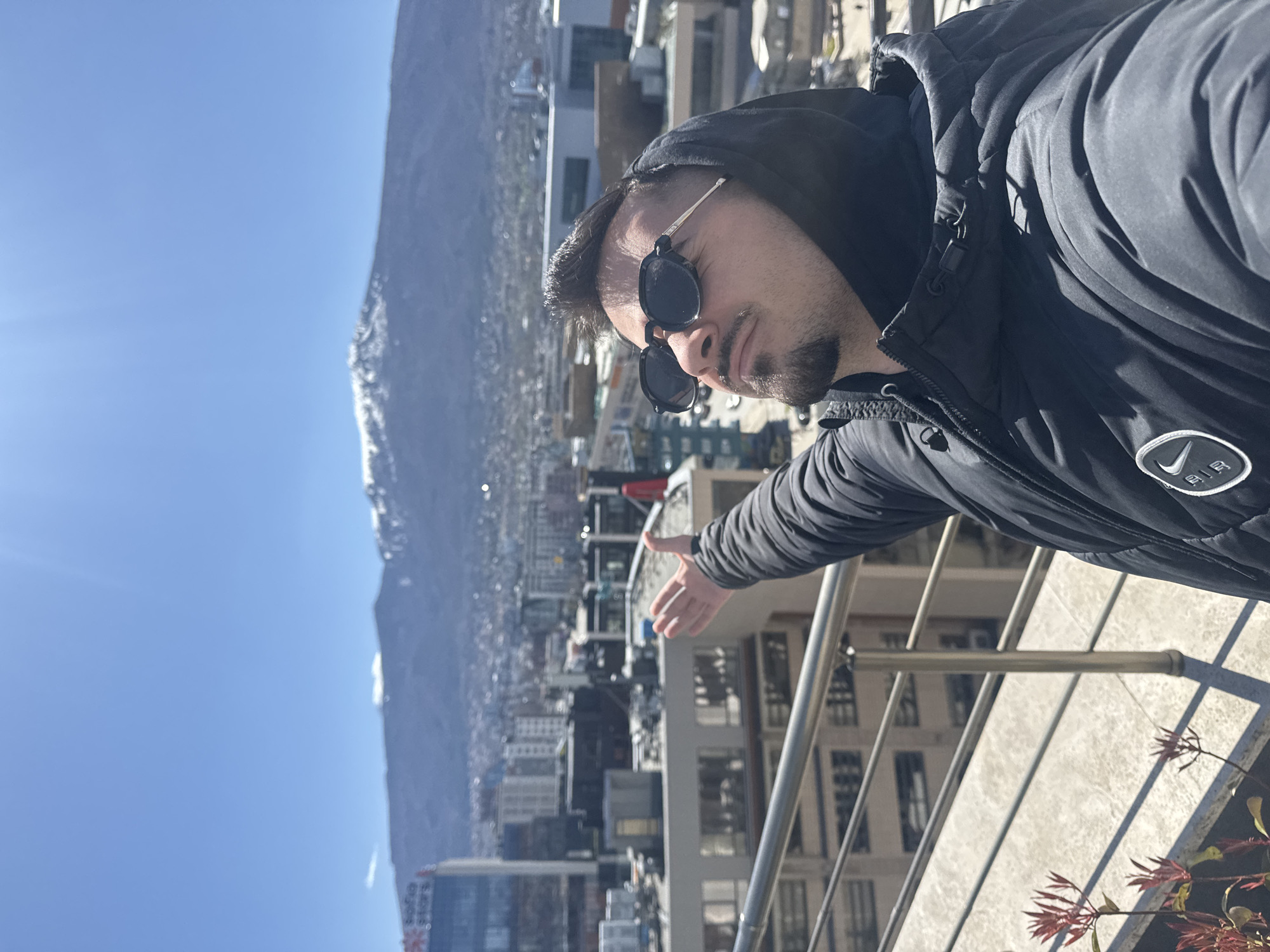 A man in a black hooded jacket and sunglasses stands on a rooftop, extending his arm towards the cityscape of Sofia, Bulgaria. In the background, a mountain range is visible against a clear blue sky. The man is wearing a Nike jacket. A metal railing and plant are at the bottom right of the image.