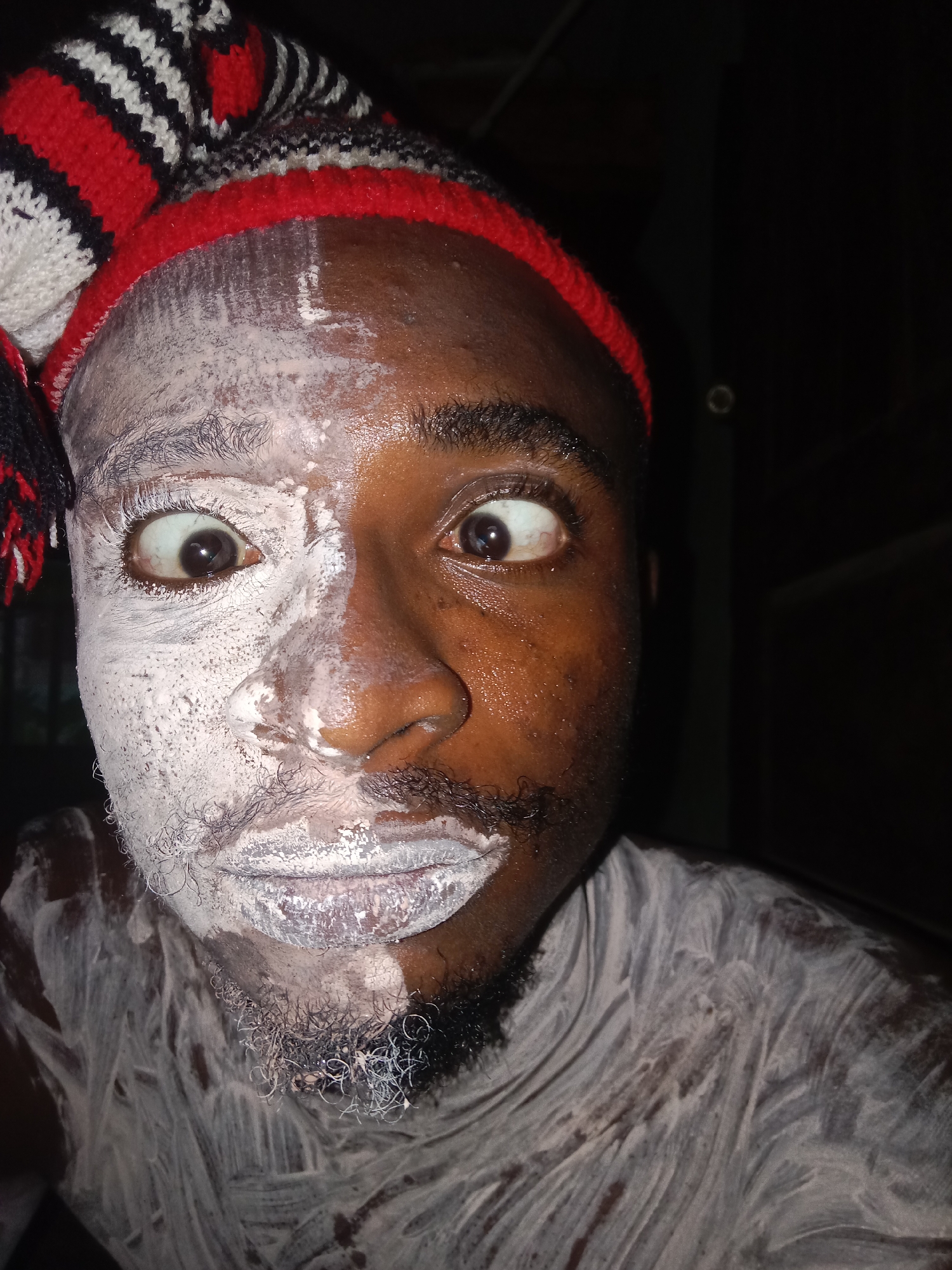 A close-up photograph captures a dark-skinned person wearing a red, white, and black knit hat. Half of the face and upper chest are covered in white cream or paste. The person's eyes are wide and appear startled. The background is dark and indistinct. The image was taken in the city of Unknown, Unknown.