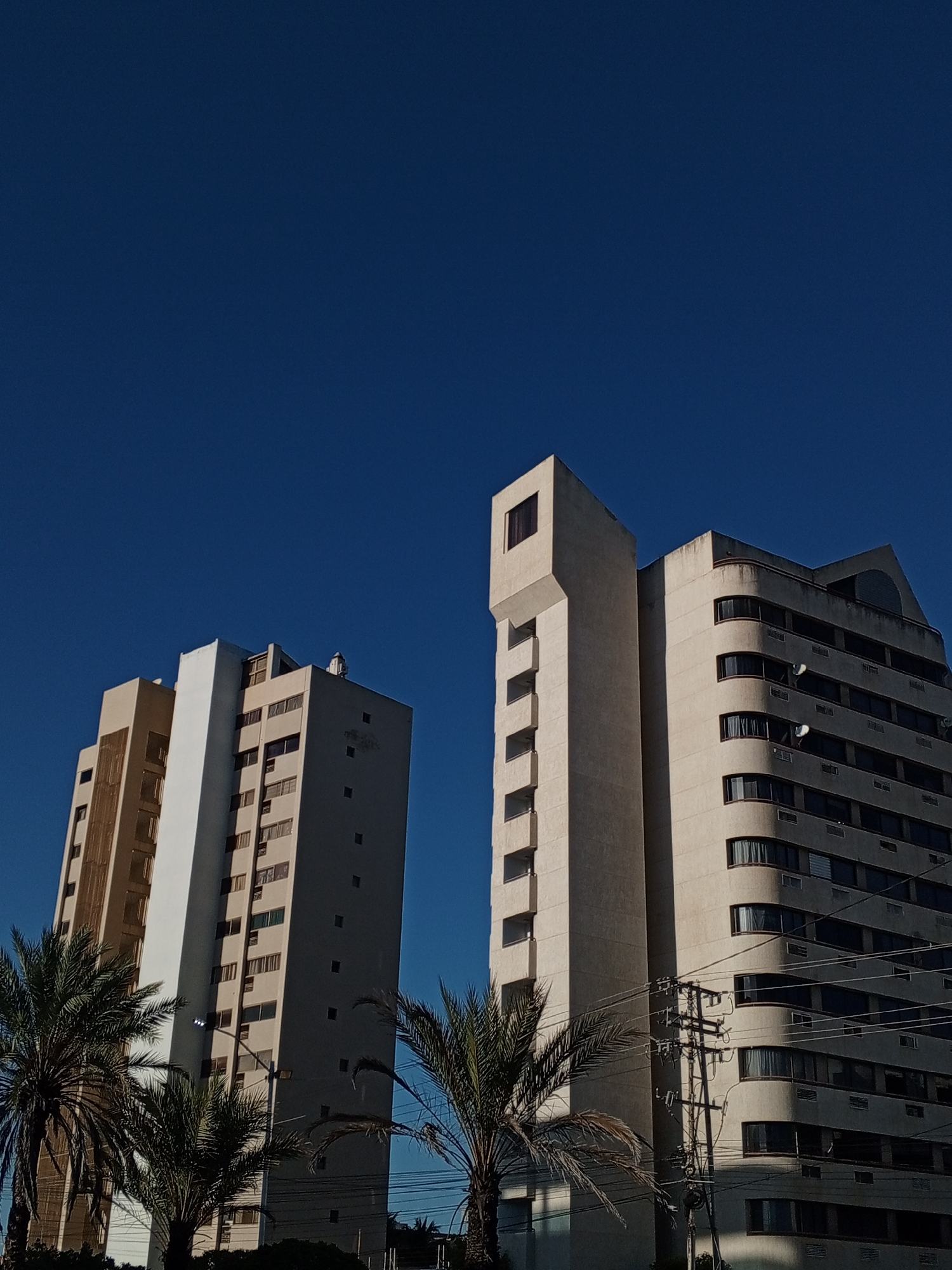 This low-angle photograph captures two prominent multi-story buildings against a striking, cloudless blue sky, characteristic of Porlamar, Venezuela. The bright daylight creates strong shadows, suggesting a mid-morning or early afternoon setting.
The building on the left is a tall structure featuring light beige and white facades with numerous rectangular windows. Its design incorporates both flat surfaces and vertical recessed sections, adding architectural detail.
The building on the right presents a more complex and aged appearance. It's also light beige but shows visible weathering and dirt, especially on its upper levels. A distinctive, angular top section with a single window crowns the structure. Below this, a segment with small, recessed square windows extends. The main body of this building features a series of curved balconies with dark-paned windows, and some satellite dishes are visible on these balconies.
In the foreground and mid-ground, several green palm trees frame the lower parts of the buildings, enhancing the tropical urban feel. Utility poles and a dense network of overhead power lines are visible, particularly in front of the right building.
The scene is static, devoid of human activity. No visible text is present in the image.