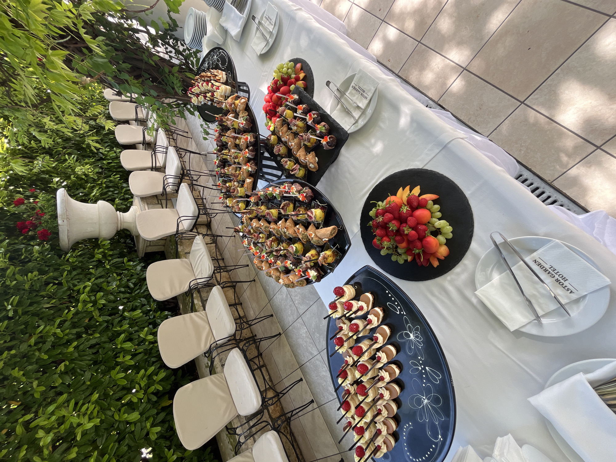 The image depicts an outdoor catering setup on a tiled patio at the Astor Garden Hotel in St. Constantine and Helena Resort, Varna, Bulgaria. A long table draped with a white tablecloth extends from the foreground towards the background, laden with various food platters.
In the foreground, on the table's left side, a black elongated platter holds multiple individual desserts, each topped with a raspberry and a chocolate stick. To its right, a round black platter features a variety of small savory bites on skewers and in cups, alongside what appear to be mini cannoli. Further right, another black, slate-like platter displays more appetizers, including items wrapped in prosciutto and various small pastries. A circular black platter in the bottom-left of the table presents an arrangement of fresh fruit, including strawberries, sliced peaches or apricots, and green grapes.
Stacks of white ceramic plates, white napkins (some folded, some unfolded), and silver serving tongs are placed intermittently along the table. Several napkins clearly bear the "ASTOR GARDEN HOTEL" logo in black text.
The background reveals a row of approximately eight to nine empty outdoor chairs with black wrought iron frames and light cream-colored cushions, arranged facing the table. Behind these chairs is a dense, lush green hedge. Towards the left of the background, a large, ornate white stone urn containing red flowers (possibly roses) is partially visible within the greenery. A mature tree with climbing vines is positioned on the right, casting dappled shadows onto the light-colored tiled patio floor. The scene is illuminated by bright sunlight, suggesting an daytime outdoor event. No people are visible, and no actions or interactions are depicted.