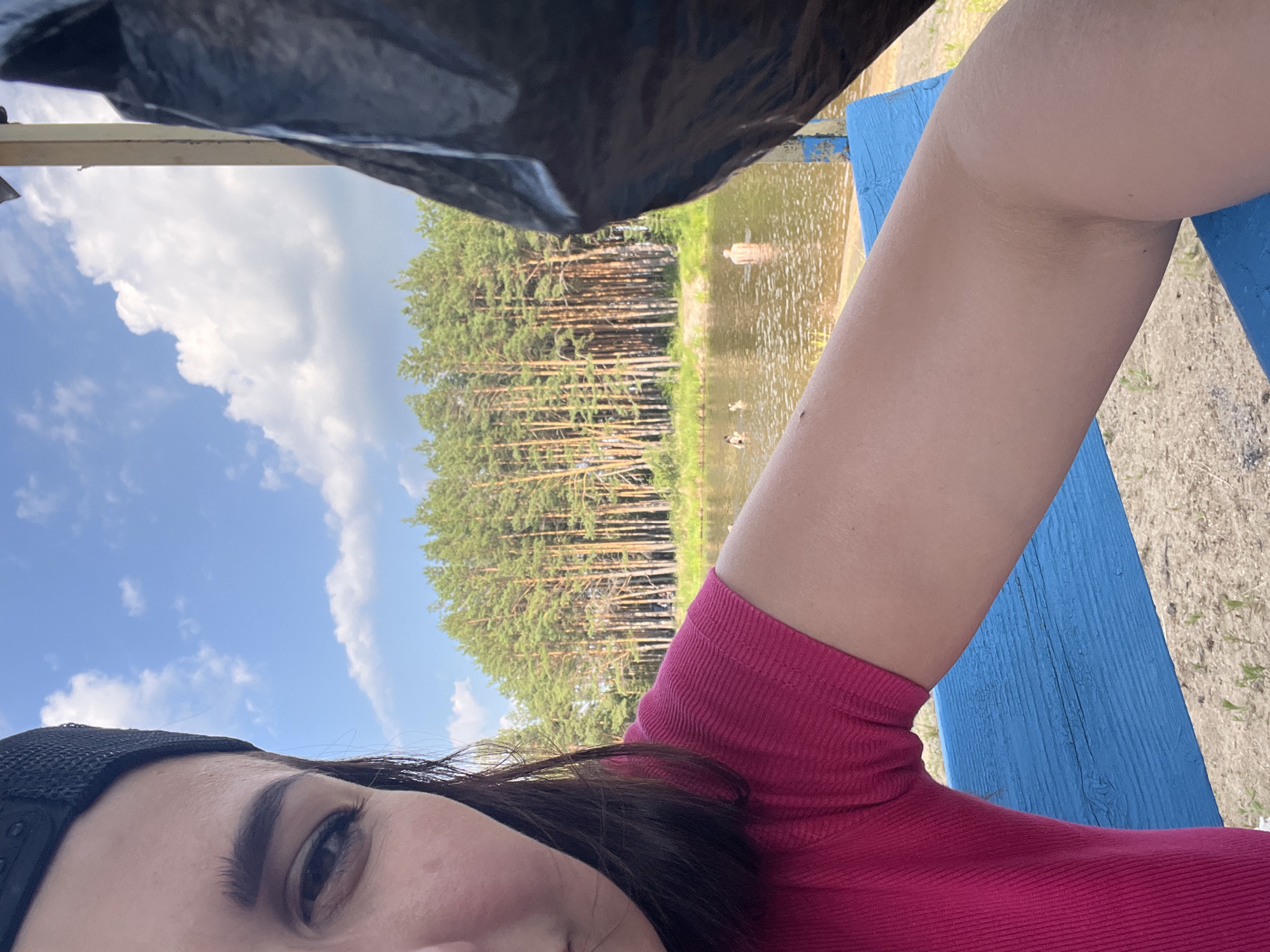 A half-face, close-up selfie of a woman wearing a black baseball cap and a bright pink, short-sleeved, ribbed shirt is in the foreground. She is looking towards the left of the frame. Her left arm, tanned, is extended towards the right. In the middle ground, a body of water, likely a lake or river, is visible, with a few people swimming. The background consists of a dense forest of tall pine trees. A blue wooden bench is in the lower portion of the image. A large black object, possibly a bag or tarp, obscures the far right side of the frame. The sky is blue with scattered white clouds. The overall scene suggests an outdoor recreational setting, possibly a park or beach area.