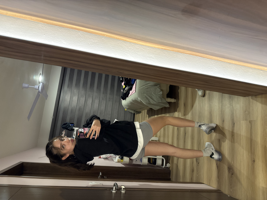 A woman stands in a doorway, holding a phone up for a mirror selfie. She wears a black sweatshirt, gray shorts, socks, and gray sneakers. A bed with a green comforter is visible behind her, along with what appears to be a rack of cleaning supplies. The room has wood-look flooring and dark blinds. A white ceiling fan is present in the upper left corner. The doorway trim is light-colored.
