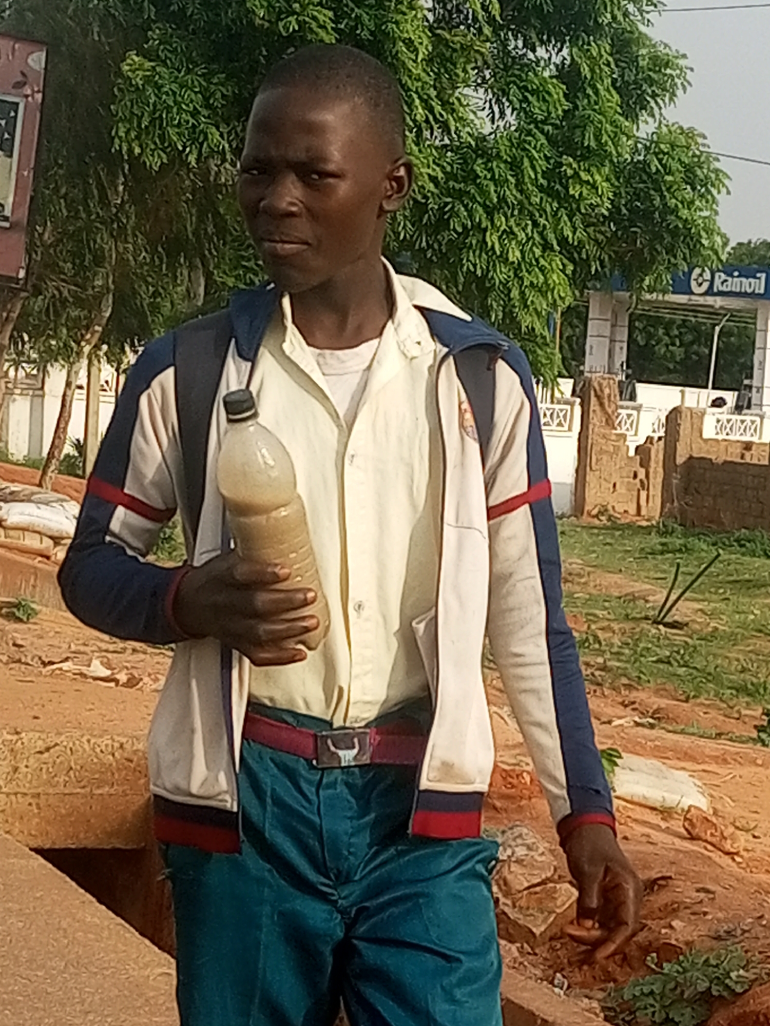 In this medium shot, an African boy, appearing to be in his late teens, stands outdoors, facing the camera. He is holding a clear plastic bottle filled with a light brown liquid in his right hand. He is wearing a white collared shirt, a jacket with blue, white, and red accents, a red belt with a buckle that has a bull's head design, and teal-colored pants. The background consists of greenery from trees and bushes, some piles of white sacks, and a structure with the "Rainoil" logo. The ground is predominantly reddish-brown dirt, with some scattered rocks and patchy grass. The overall lighting suggests it is daytime.