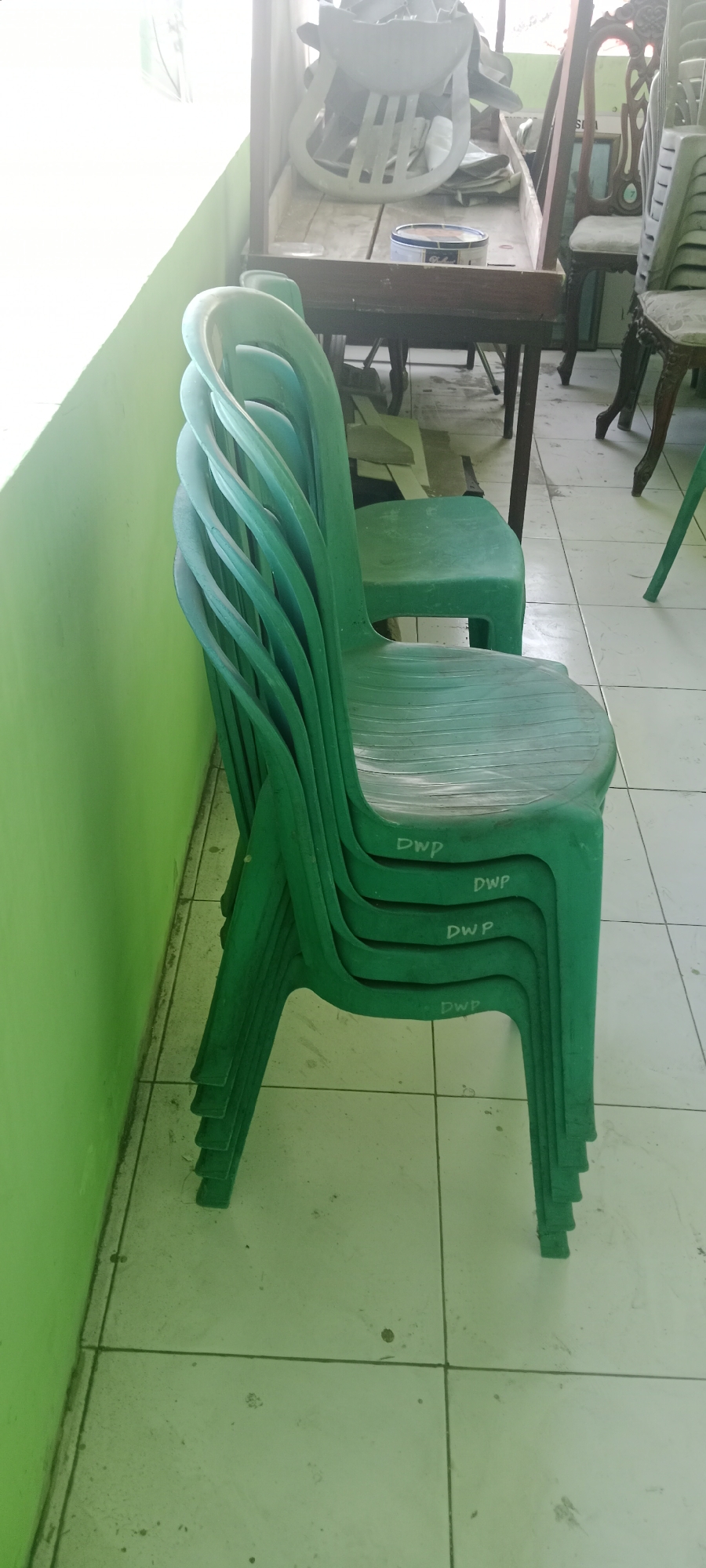 The image shows a stack of green plastic chairs in a room. The chairs have the letters "DWP" written on them. In the background, there is a wooden table with some white plastic items on it, and a few other chairs. The floor is tiled. The overall lighting suggests it is daytime, possibly indoors with natural light coming from a window or opening. The scene appears to be located in a storage area or a functional space. The text on the chairs suggests they might belong to an organization or facility in Tuban, Indonesia.
